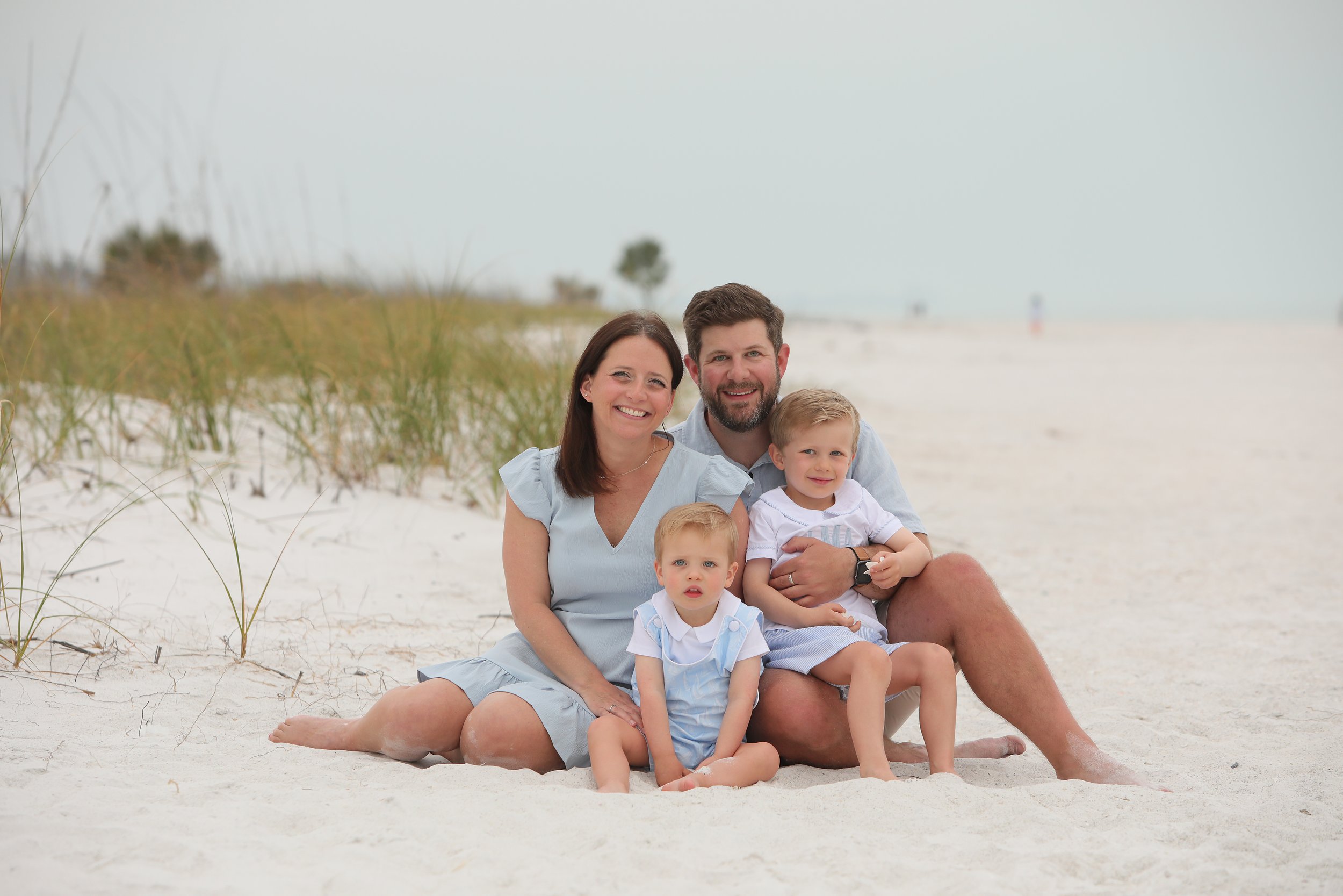 Waterrock Construction owners sitting on the sand at the beach, smiling at the camera. The mother and father are seated with their two young children. The background features beach grass.