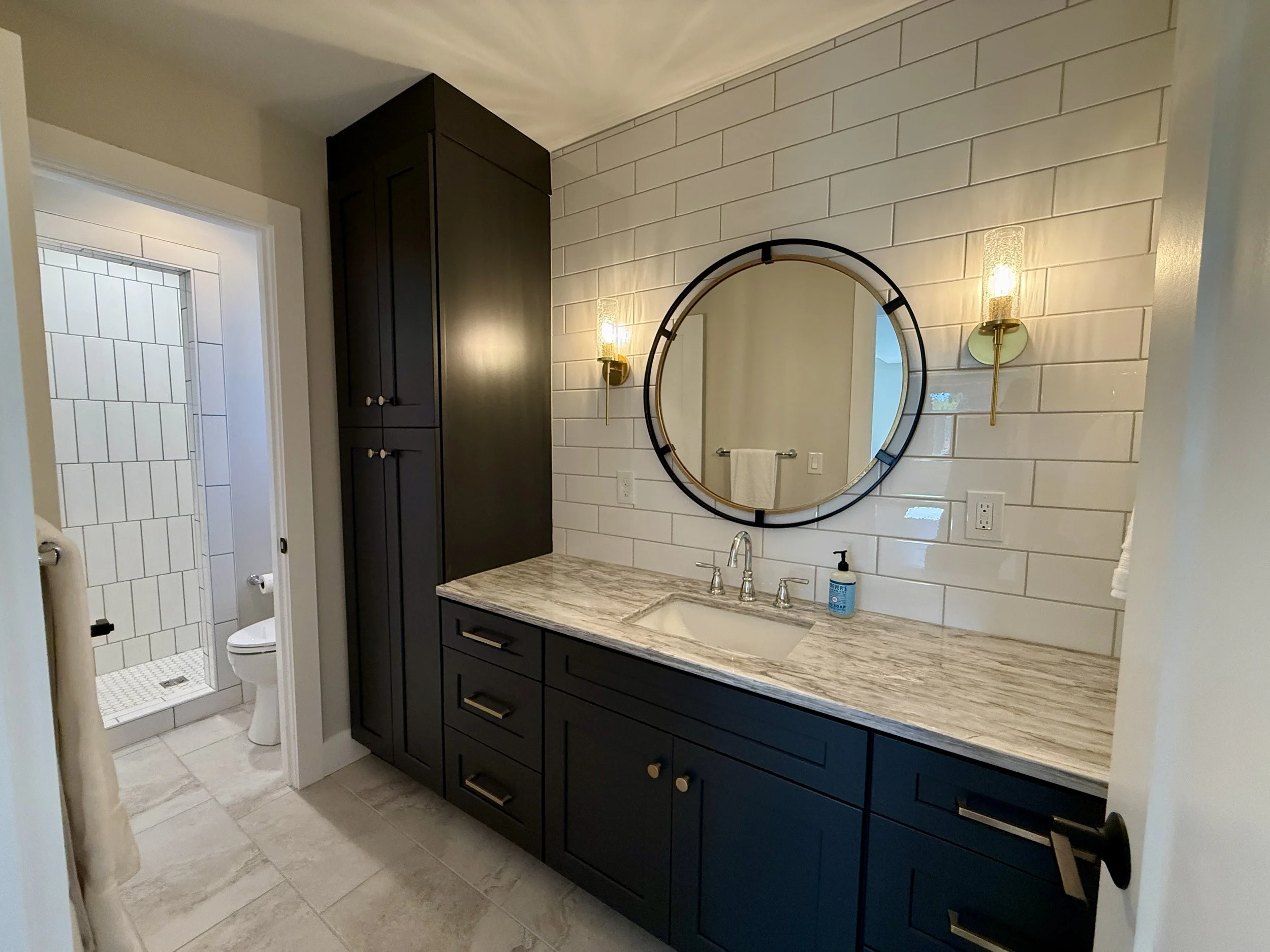 Bathroom with dark cabinetry, marble-look countertop, round mirror, and tiled shower inside a custom-built one-bedroom DADU.