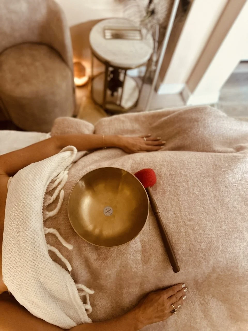A person sitting on a bed or couch, holding a brass singing bowl and a wooden mallet with a red tip, wearing cozy beige clothing. A side table with a glass top and decorative items is visible in the background.