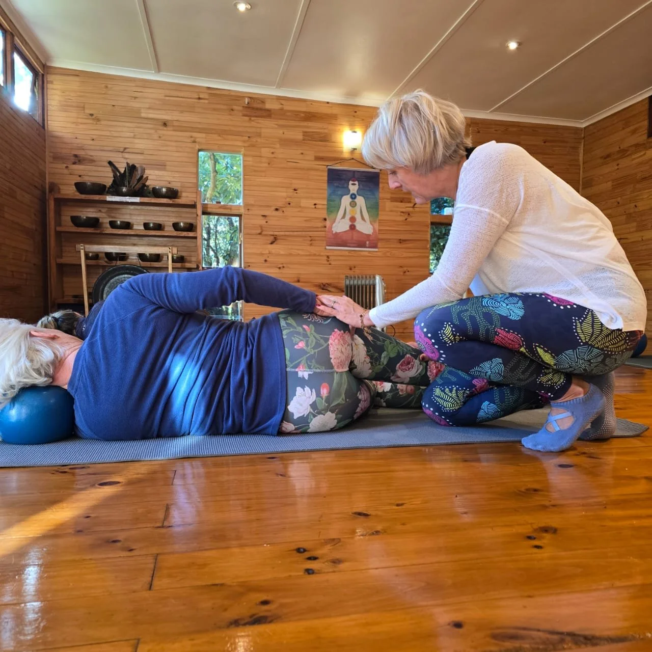 A woman lying on her side on a yoga mat with her head on a blue bolster, receiving a gentle touch on her hips from a woman kneeling beside her in a yoga studio with wooden walls and shelves.