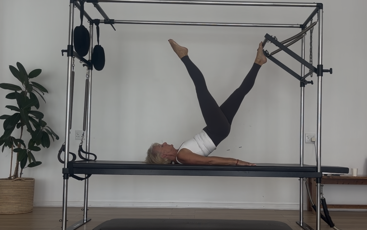 Older woman practicing Pilates on a reformer machine in a minimalistic room with a potted plant, electrical outlet, and a wooden bench