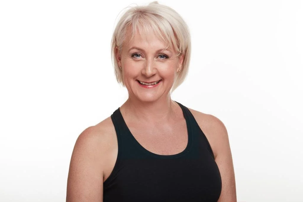A smiling middle-aged woman with short blonde hair, wearing a black athletic tank top, standing against a white background.