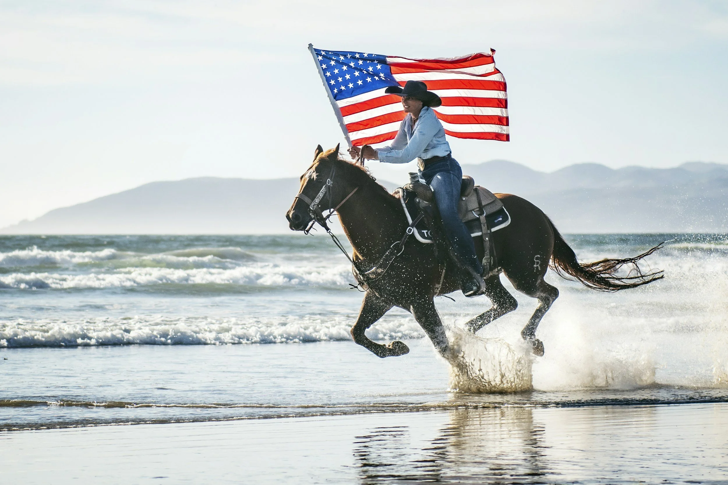 A person riding a horse on the beach while holding an American flag, with waves crashing and distant mountains in the background.