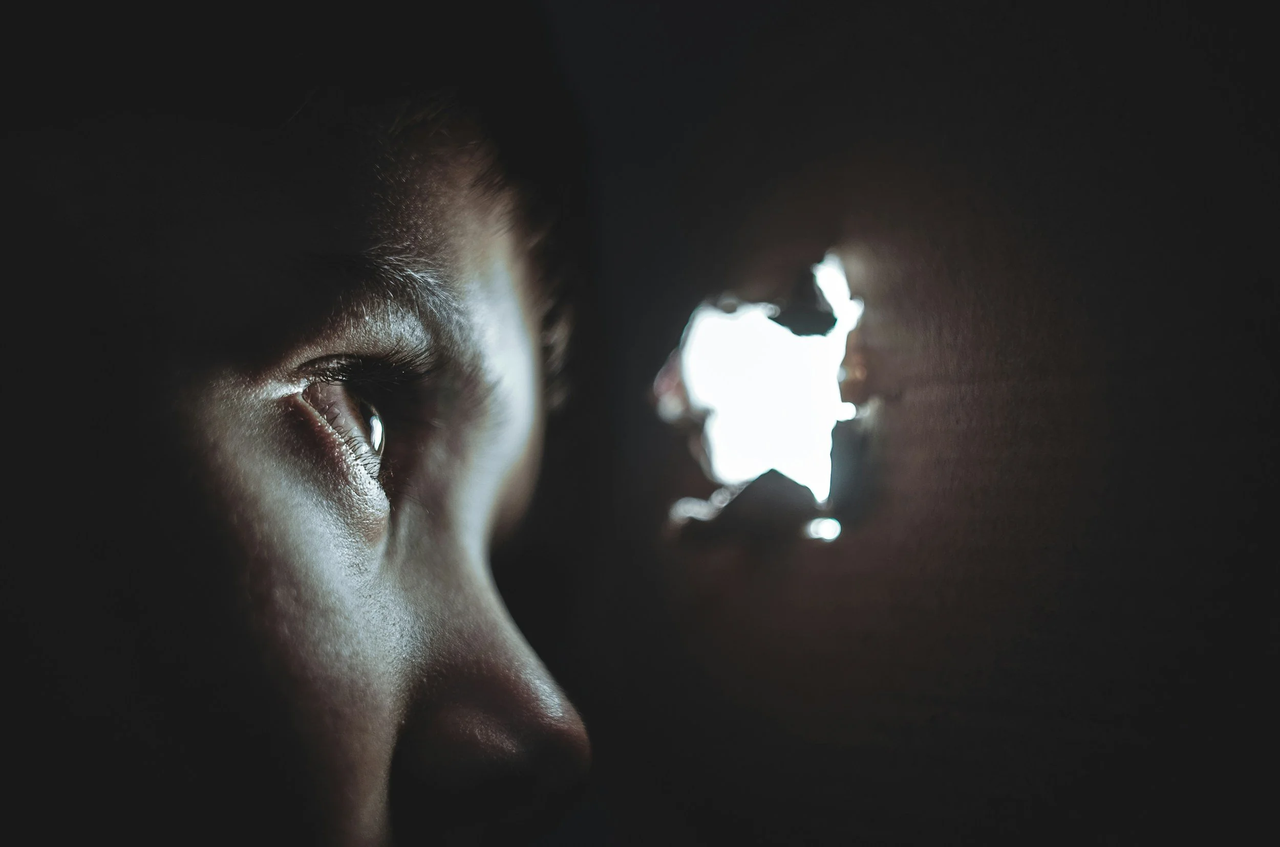 Close-up of a child's face in darkness, looking through a hole to bright light outside, with one eye visible and detailed skin texture.