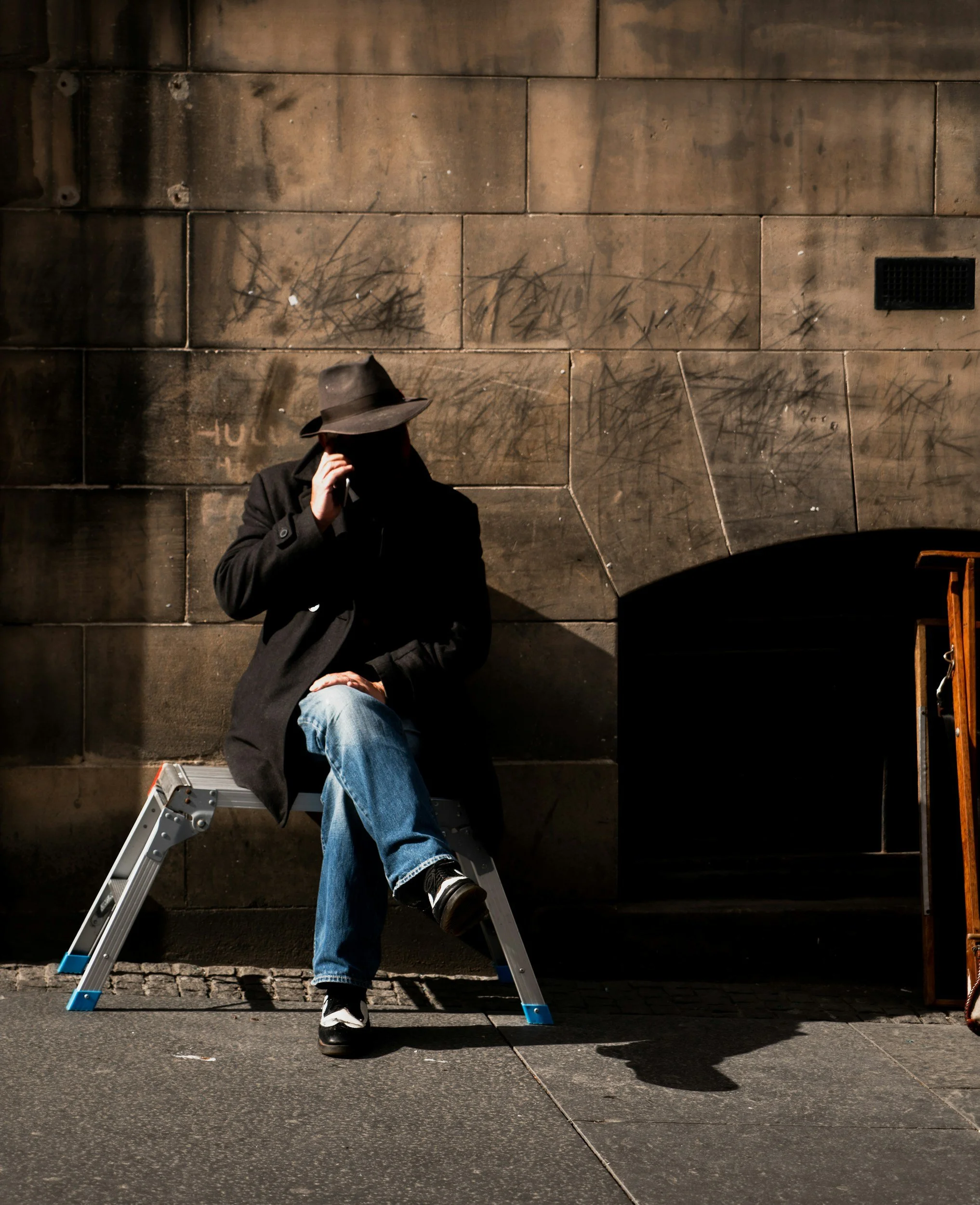 Person wearing a dark hat and coat sitting on a step ladder against a brick wall, with the person's face partially hidden and their hand near their face, possibly talking on a phone.