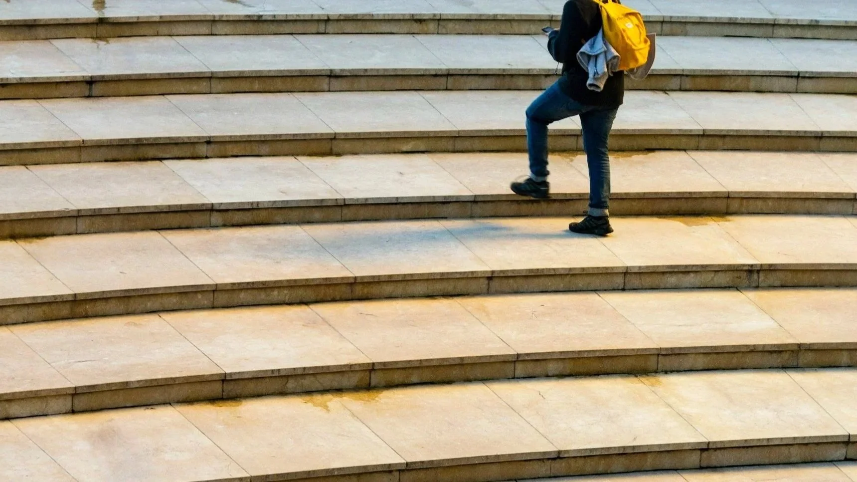 A woman wearing glasses sitting at a white desk in front of a window, working on a laptop displaying spreadsheets and graphs, with a potted plant and calculator on the desk.