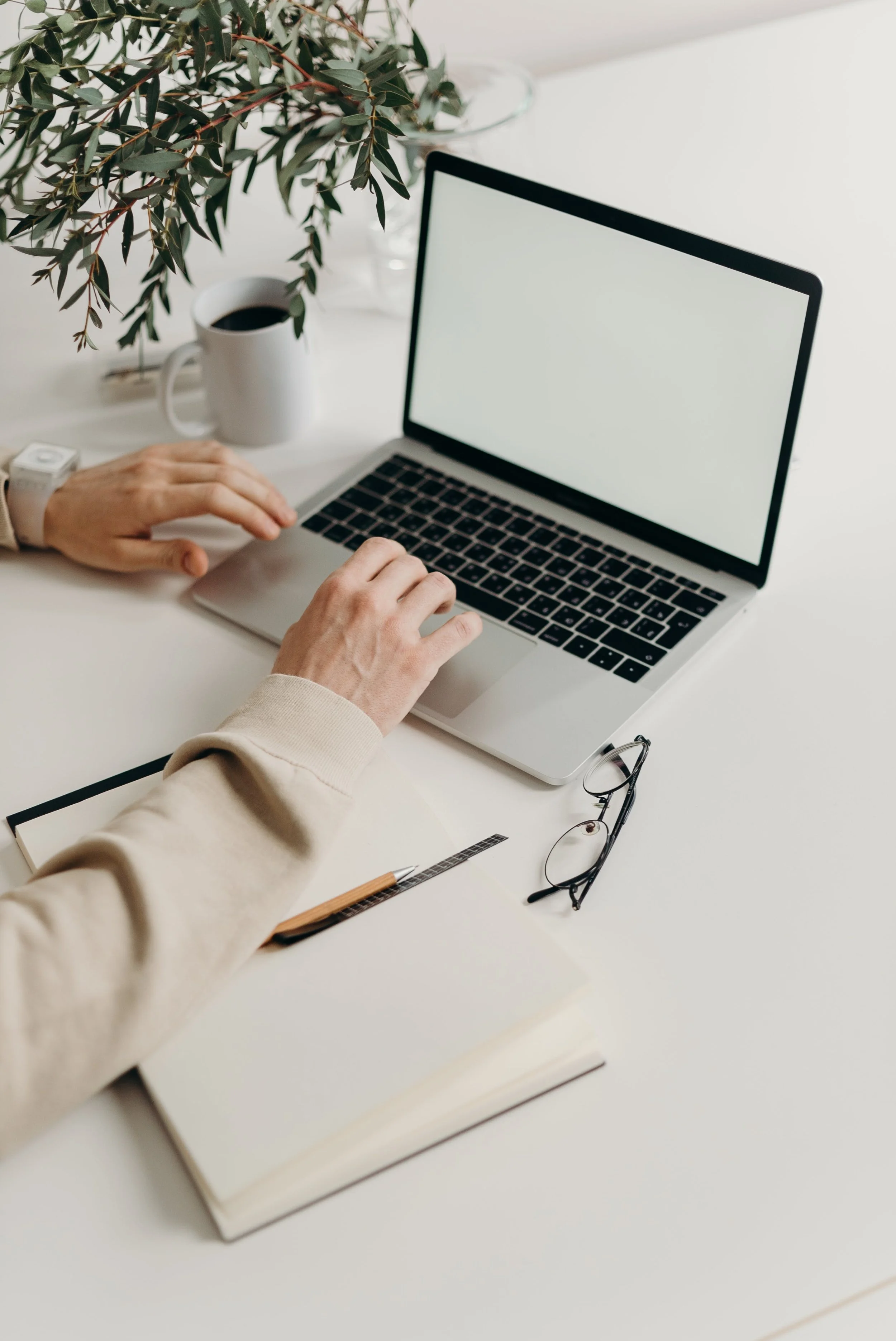 A person working on a laptop at a white desk with a pair of glasses, a closed notebook, a pen, a cup of coffee, and a plant in the background.