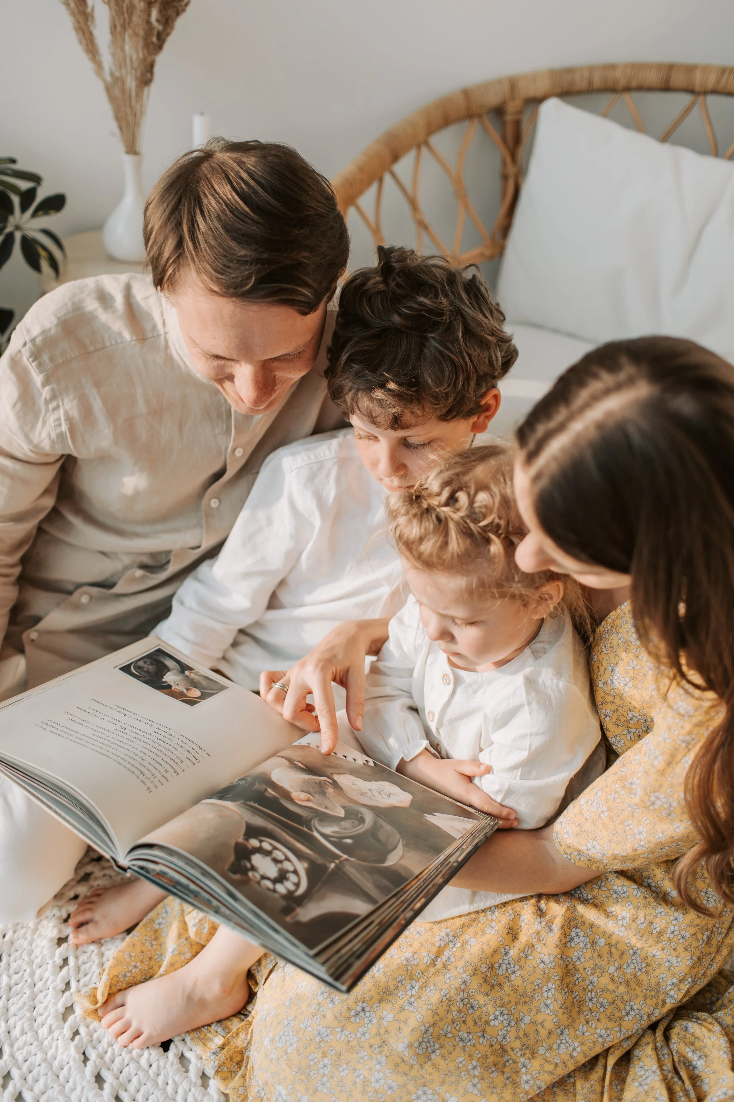 A family of five, including two children and two adults, sitting on a bed and looking at a photo album together.