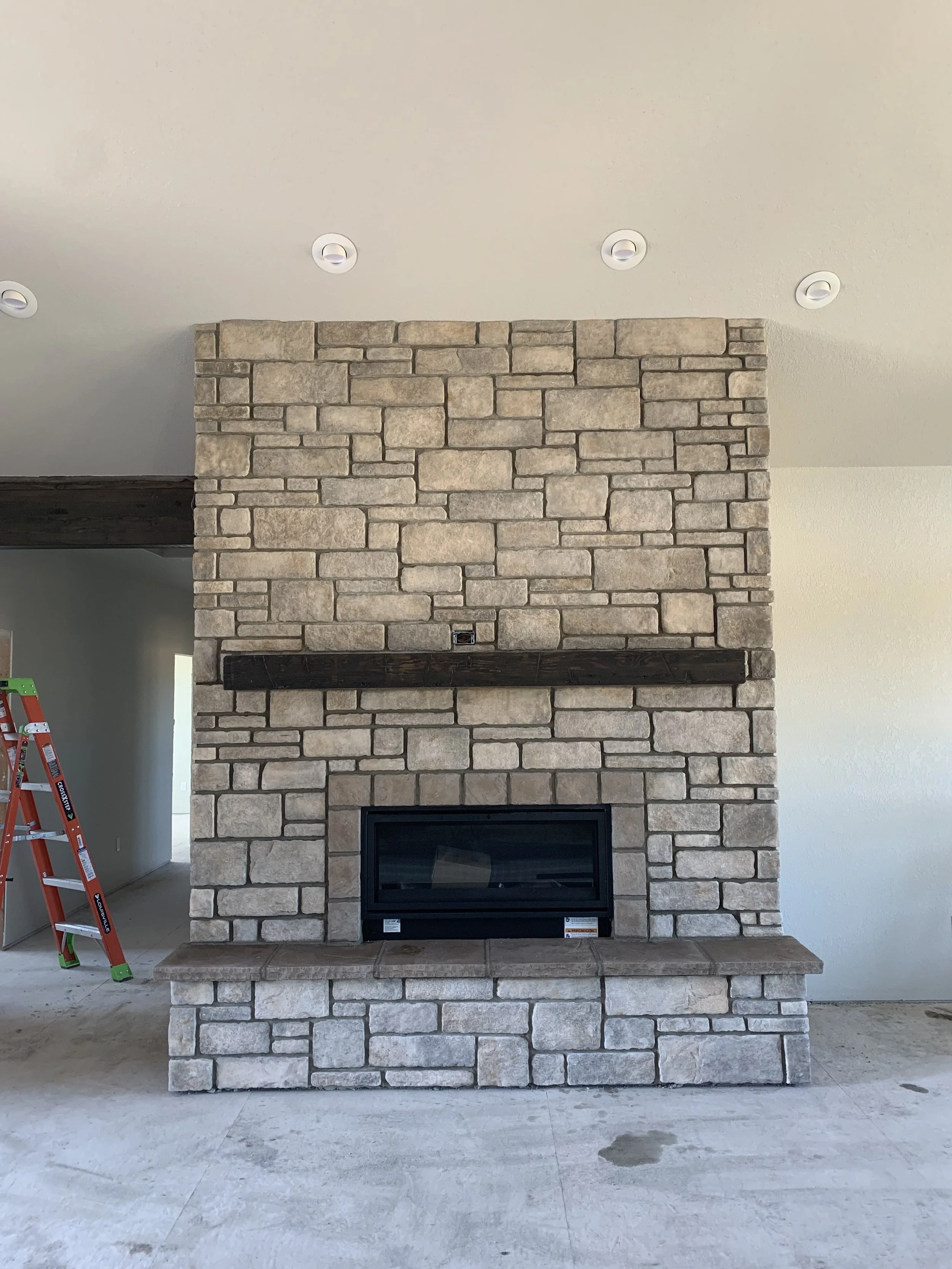 A stone fireplace with a dark wooden mantel in a room under construction, with a red ladder on the left side and unfinished flooring.
