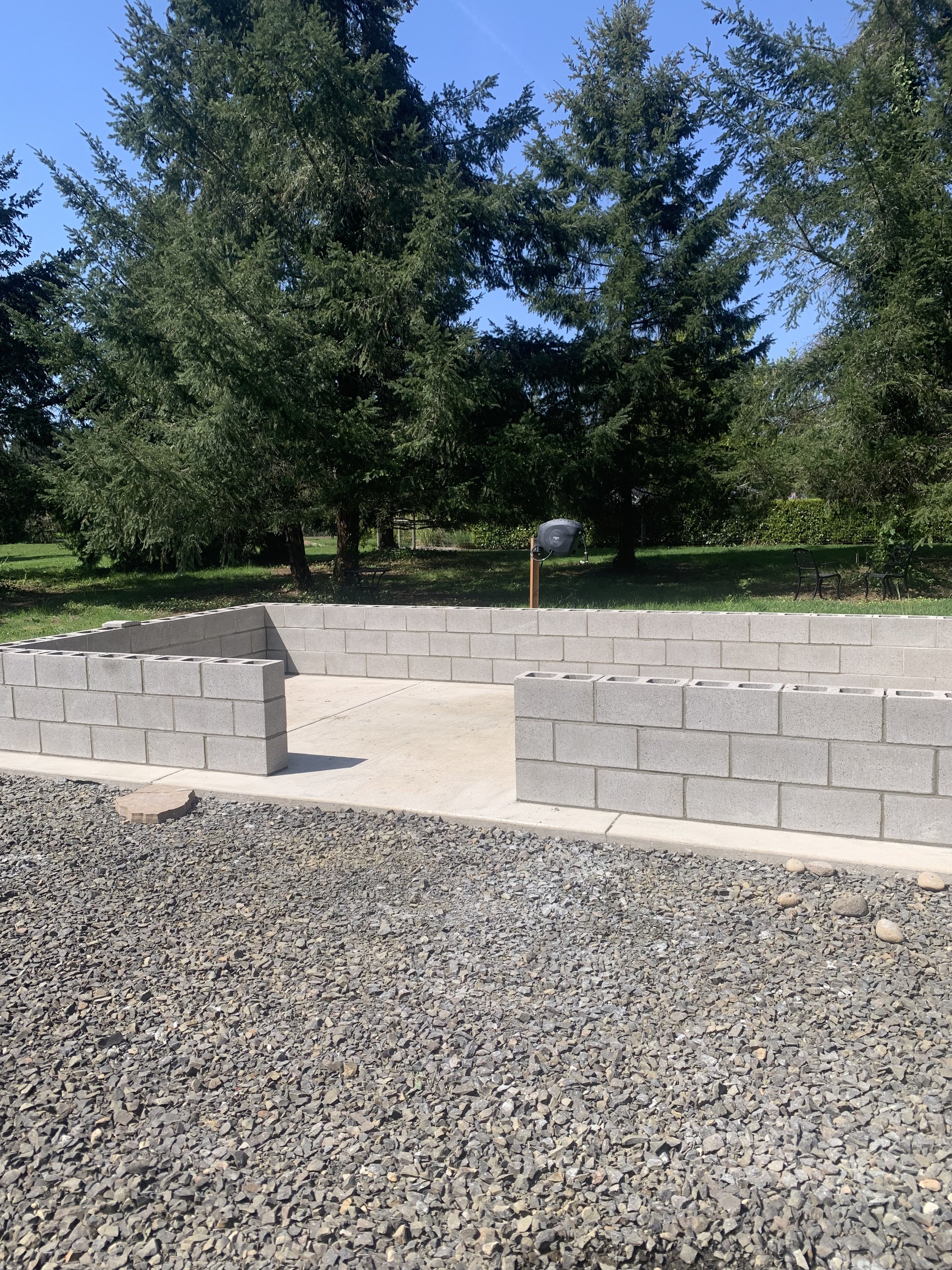 Partially built cinder block structure in an outdoor park area with trees and benches in the background, and a gravel surface in the foreground.