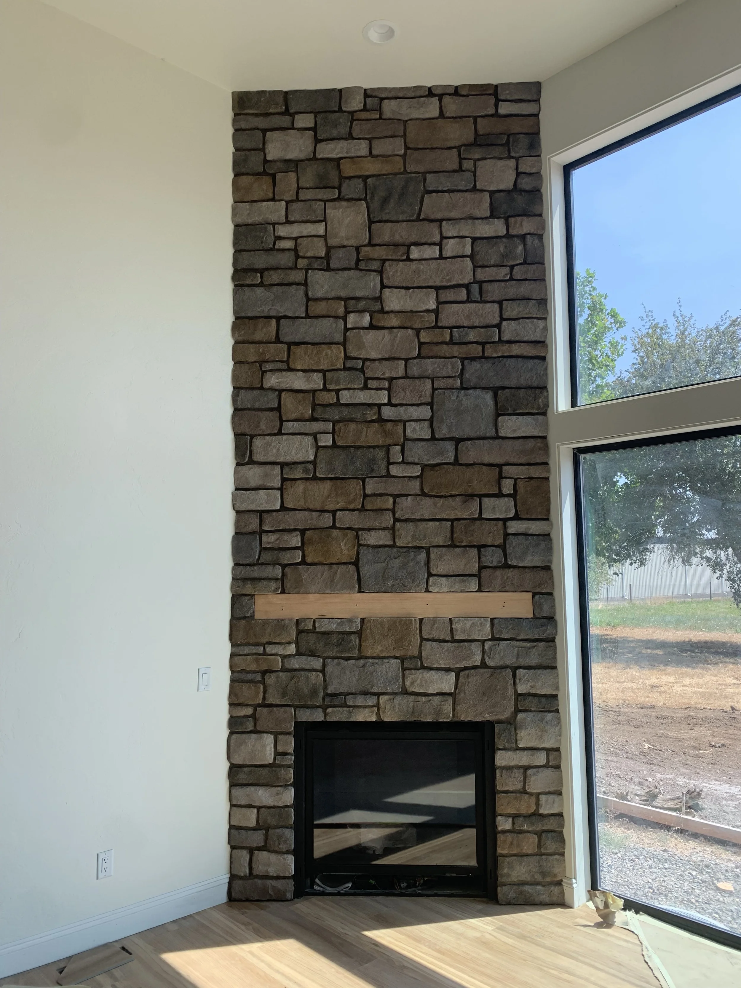 A living room with a tall stone fireplace and large window showing an outdoor yard and trees.