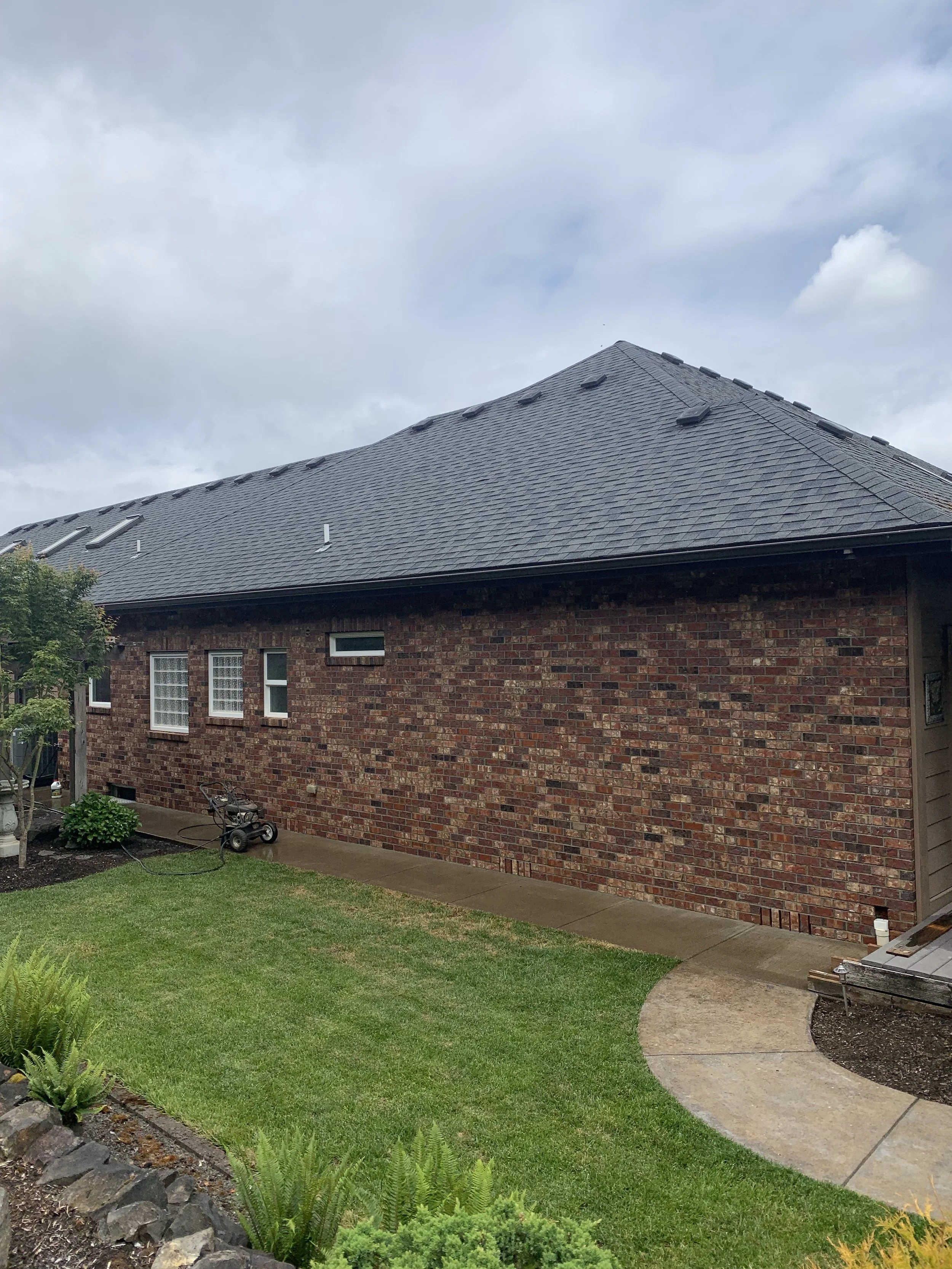 Backyard of a brick house with a curved concrete pathway, green grass, small plants, and a lawn mower near the house wall under a cloudy sky.
