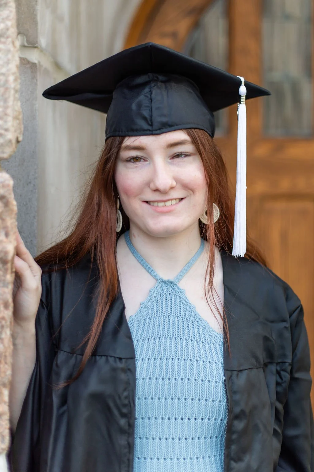 Louise Irpino, a white woman with red hair, wears a blue dress and a black cap and gown, while standing against a wall and smiling.