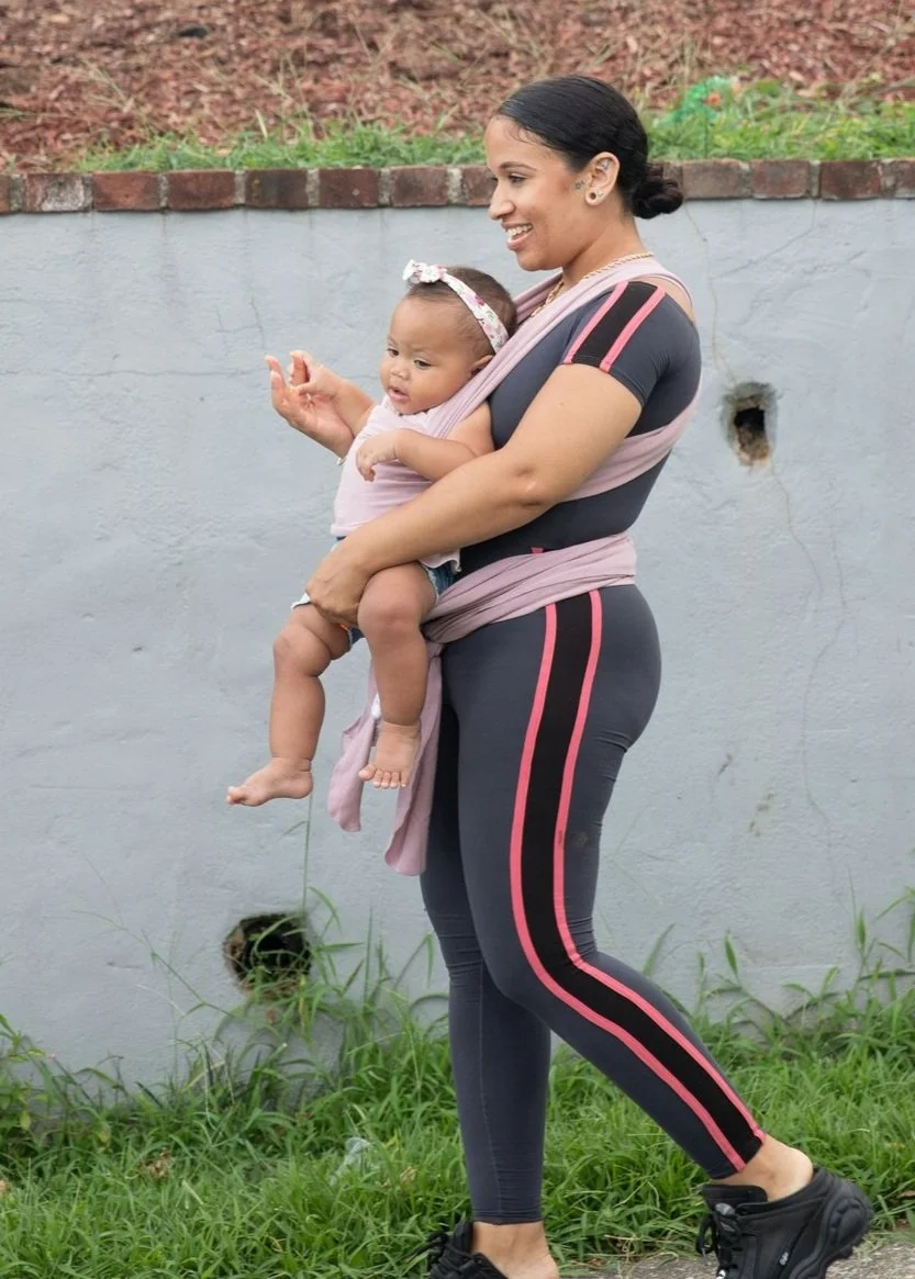 A woman in athletic clothing, smiling, carries a baby in a wrap carrier. The baby is wearing a headband and the woman is walking outdoors.