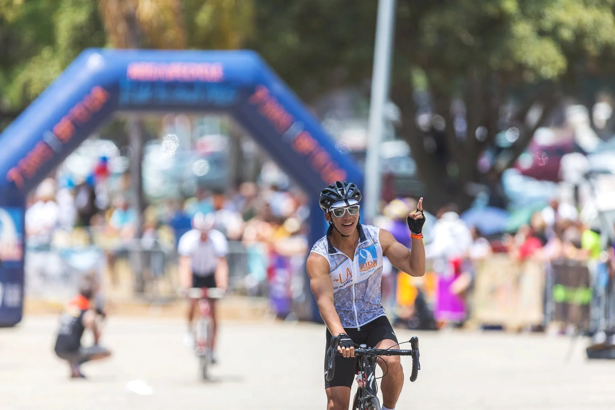 A woman riding a bicycle and smiling at a daytime outdoor event. She is wearing sunglasses, a helmet, and a athletic vest. The background has a crowd of people and an inflatable arch.