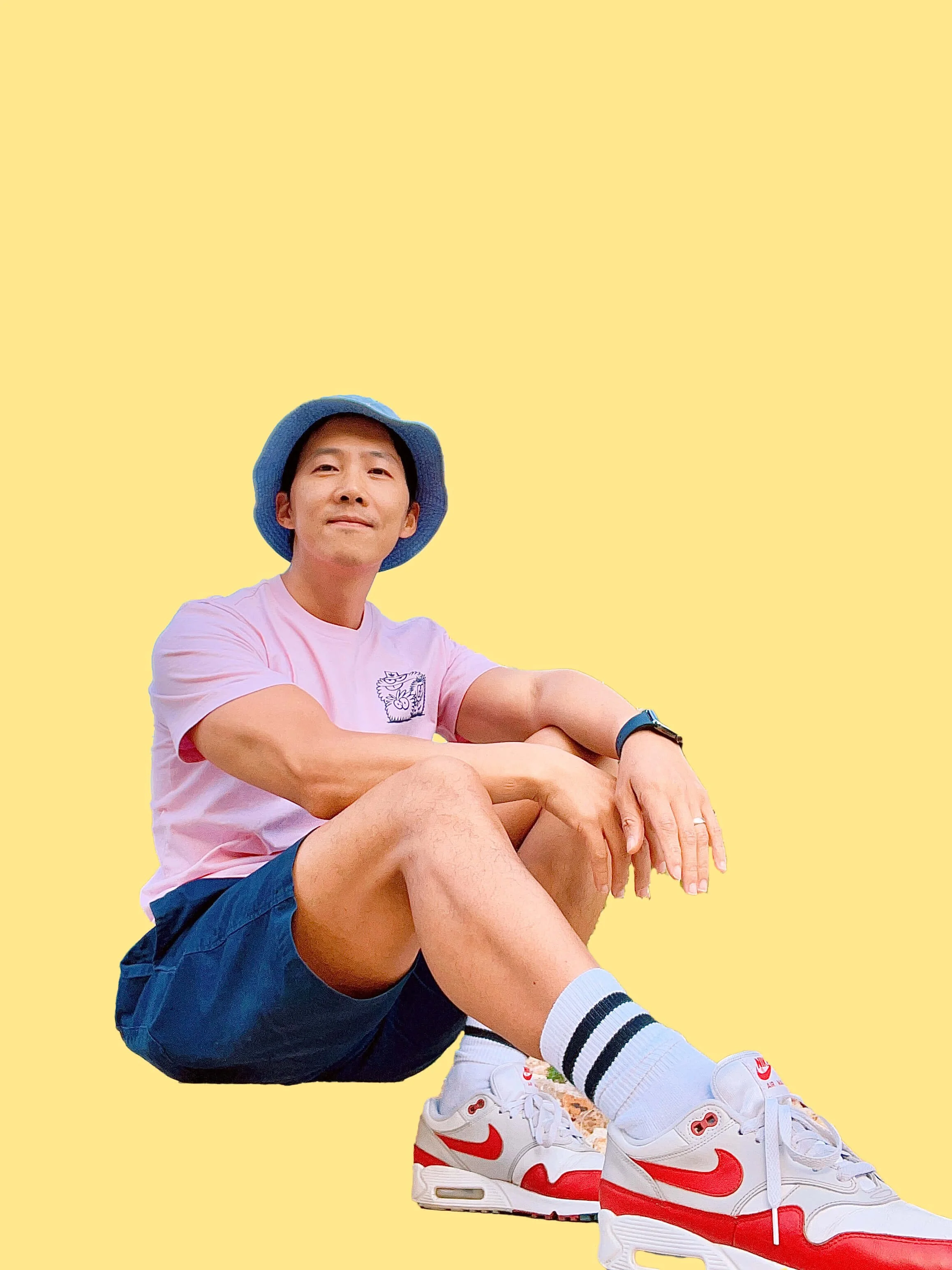 A young man sitting with his knees up, wearing a blue bucket hat, pink T-shirt with a graphic, navy shorts, white Nike sneakers with red accents, and striped socks, set against a plain yellow background.