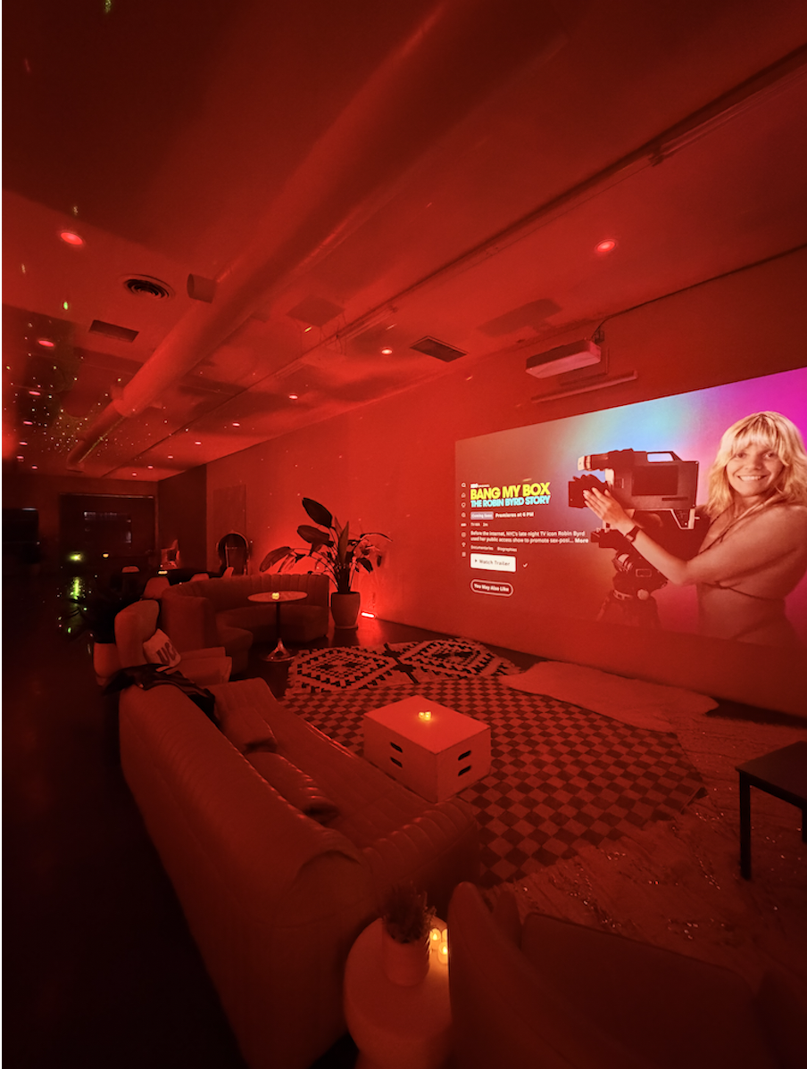 A dimly lit room with red lighting, featuring a large projection screen displaying a movie poster for "Bang My Box." The room is furnished with couches, a small potted plant, and a patterned rug, creating a cozy viewing area.