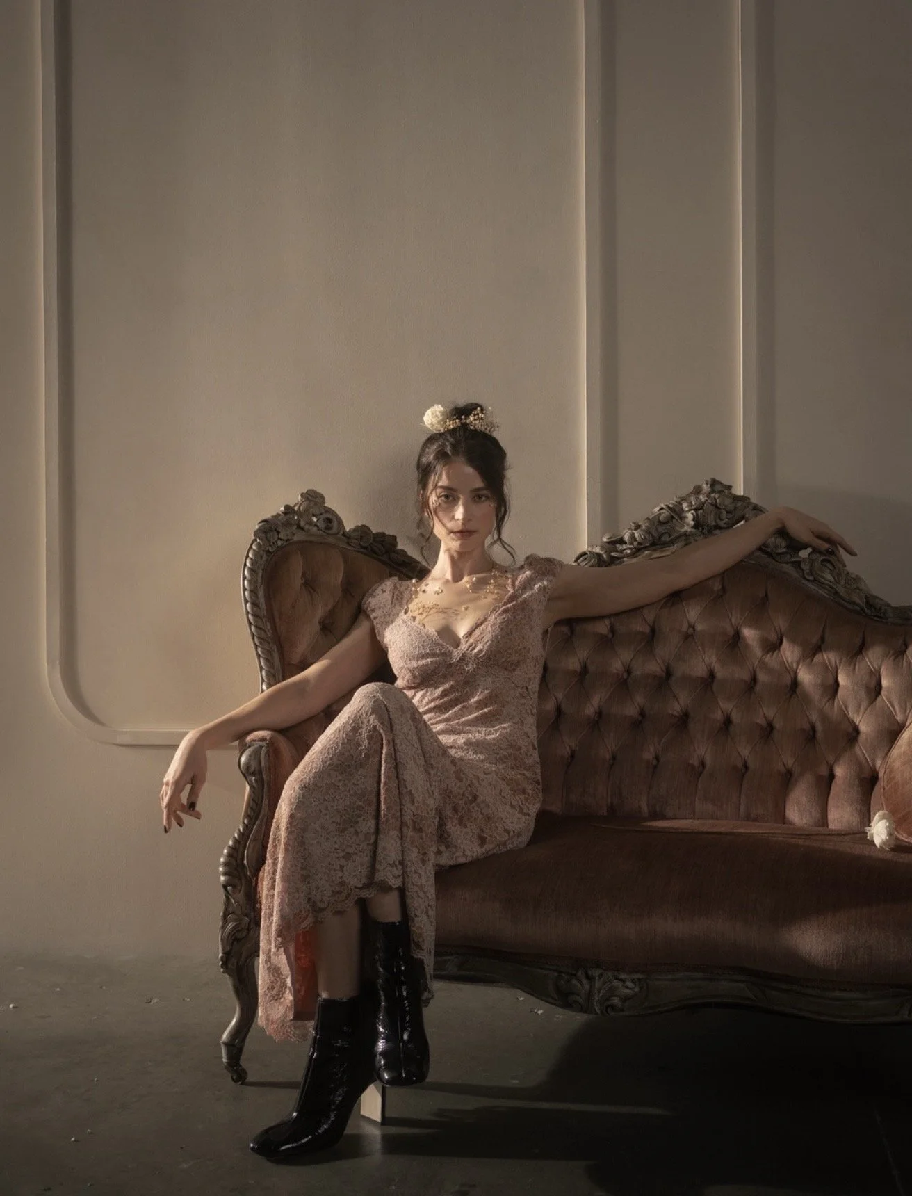 A woman in a long pink lace dress sits on an antique brown velvet sofa with ornate wood carvings, posing with one arm stretched out along the back of the sofa. She has dark hair styled in an updo with a decorative hairpiece, and is wearing black high