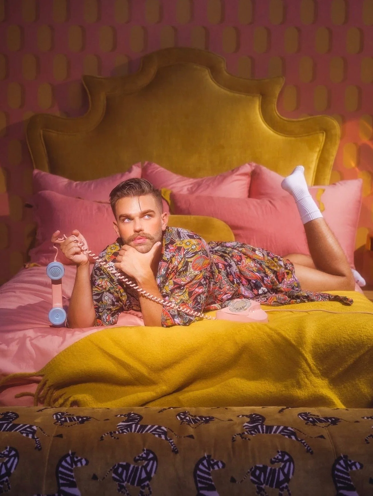 A man lying on a bed holding a retro pink rotary phone, wearing a floral shirt, with his feet up in white socks, in a colorful, vintage-style bedroom with patterned wallpaper and zebra-striped blanket.