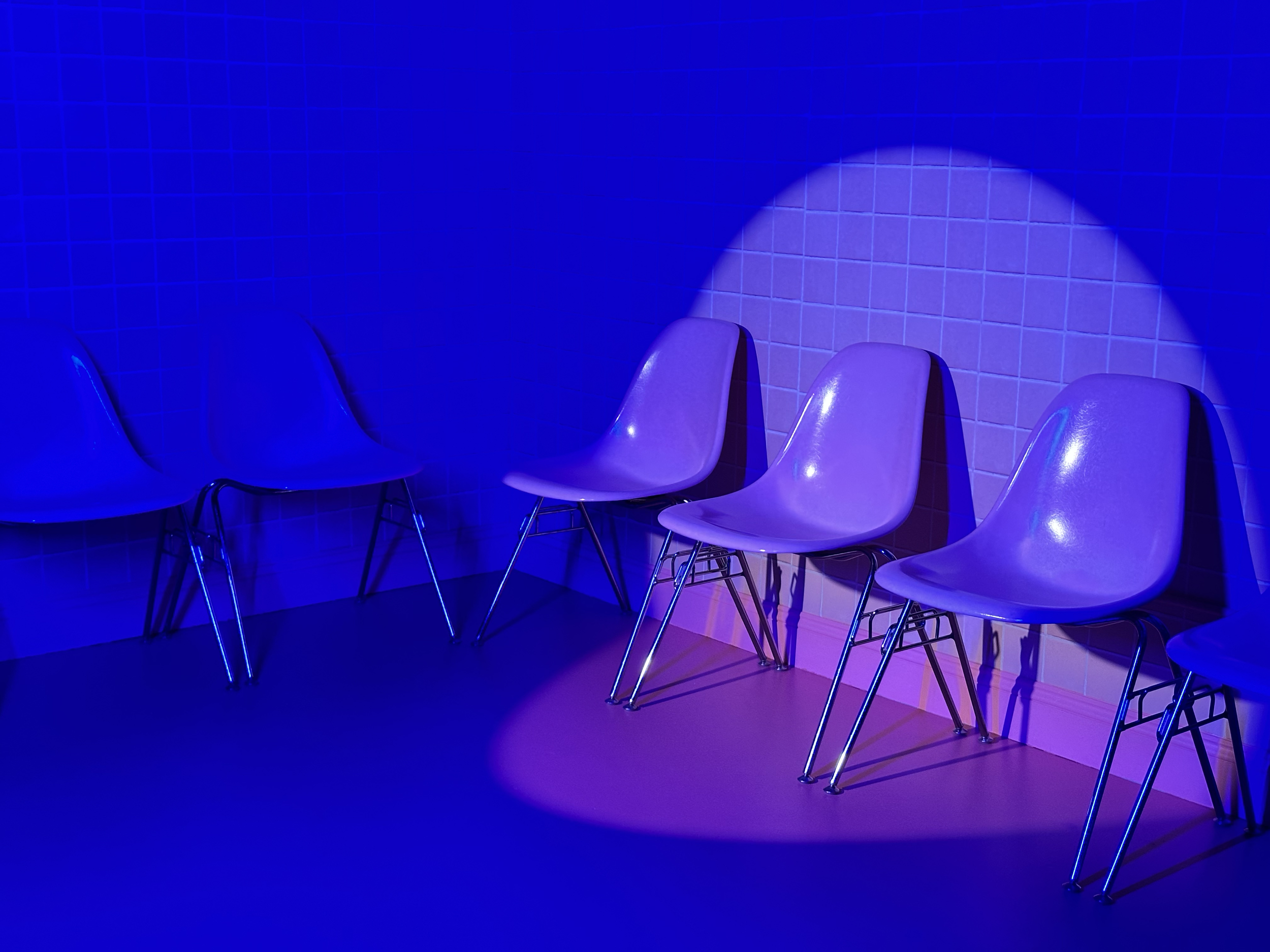 A row of white chairs with wooden legs lining a tiled wall, illuminated by a blue light in a room.