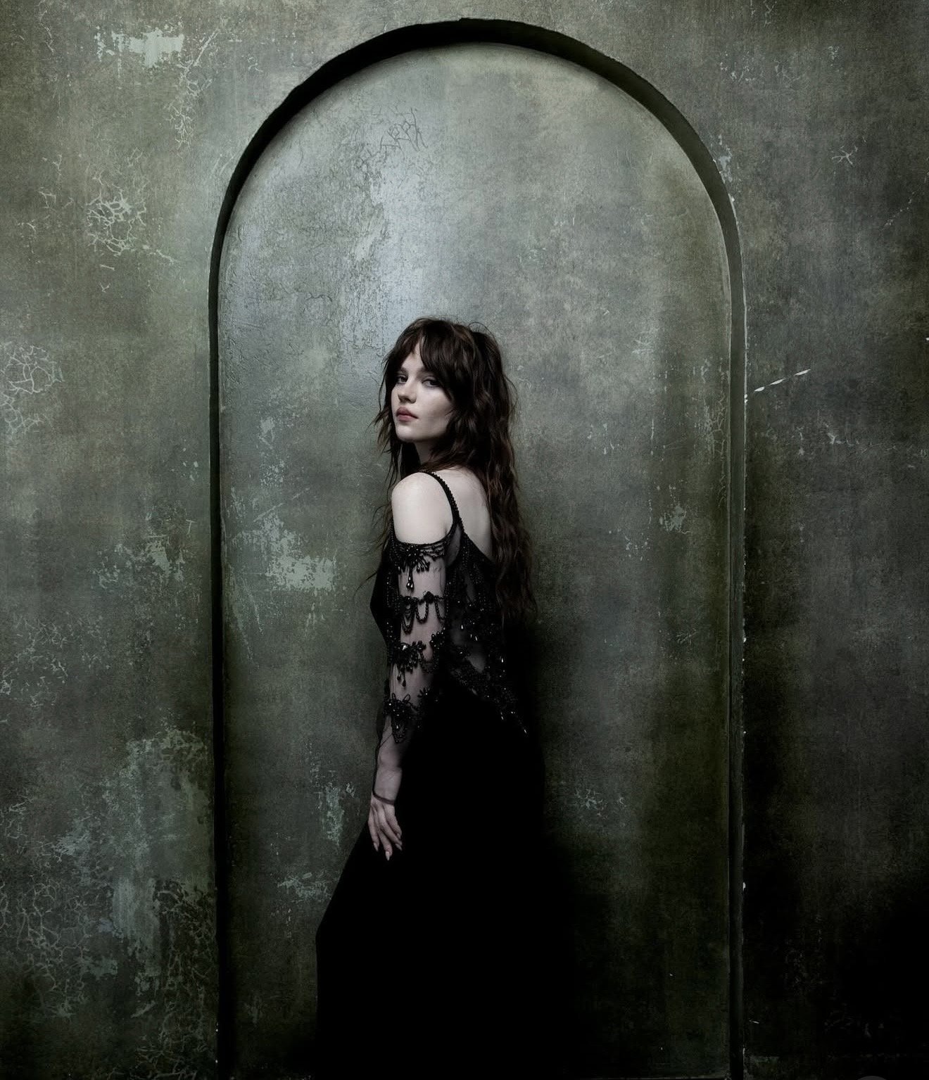 A woman with long wavy brown hair and fair skin stands against a textured, dark green wall with an arched niche. She wears a black off-shoulder dress with lace detailing and looks over her shoulder.