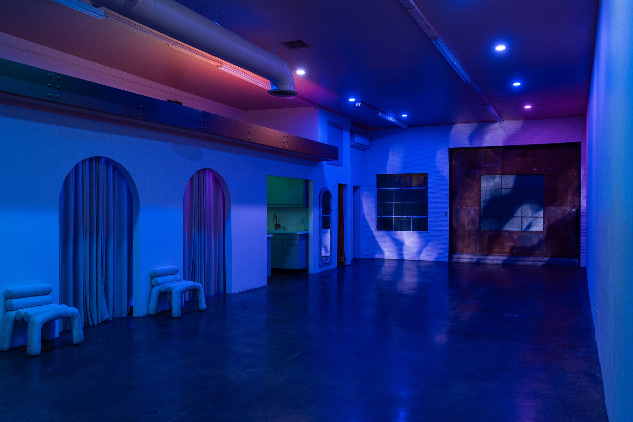 A dimly lit room with blue and purple lighting, featuring arched doorways with curtains, two modern white chairs, and a kitchenette area. The room has an industrial style with exposed ductwork and a polished concrete floor.