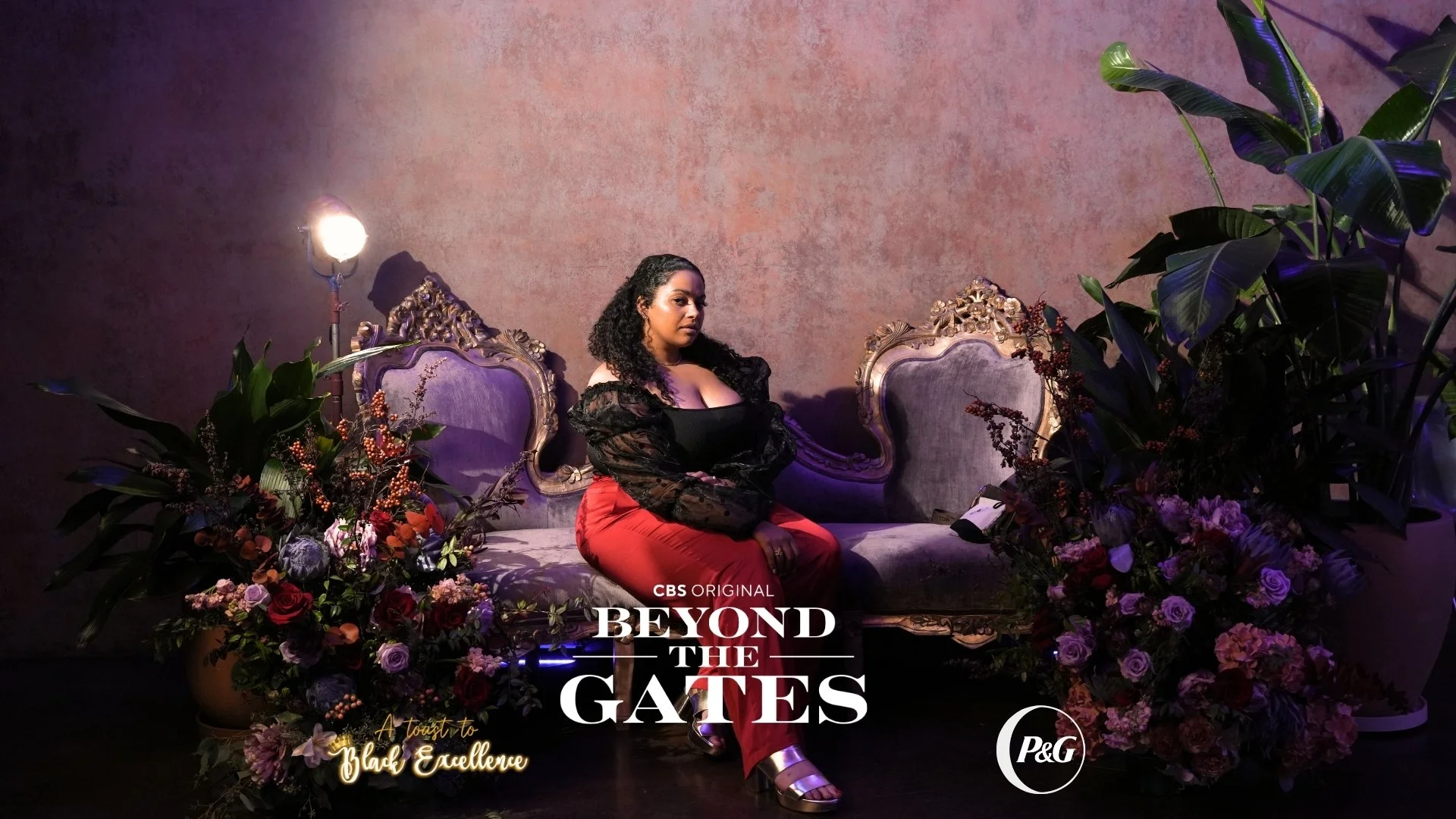 A woman with curly dark hair sitting on a vintage purple velvet sofa with ornate wood carvings, surrounded by large floral arrangements, with a pink textured wall behind her and soft lighting. The image promotes the CBS original series 'Beyond the Ga