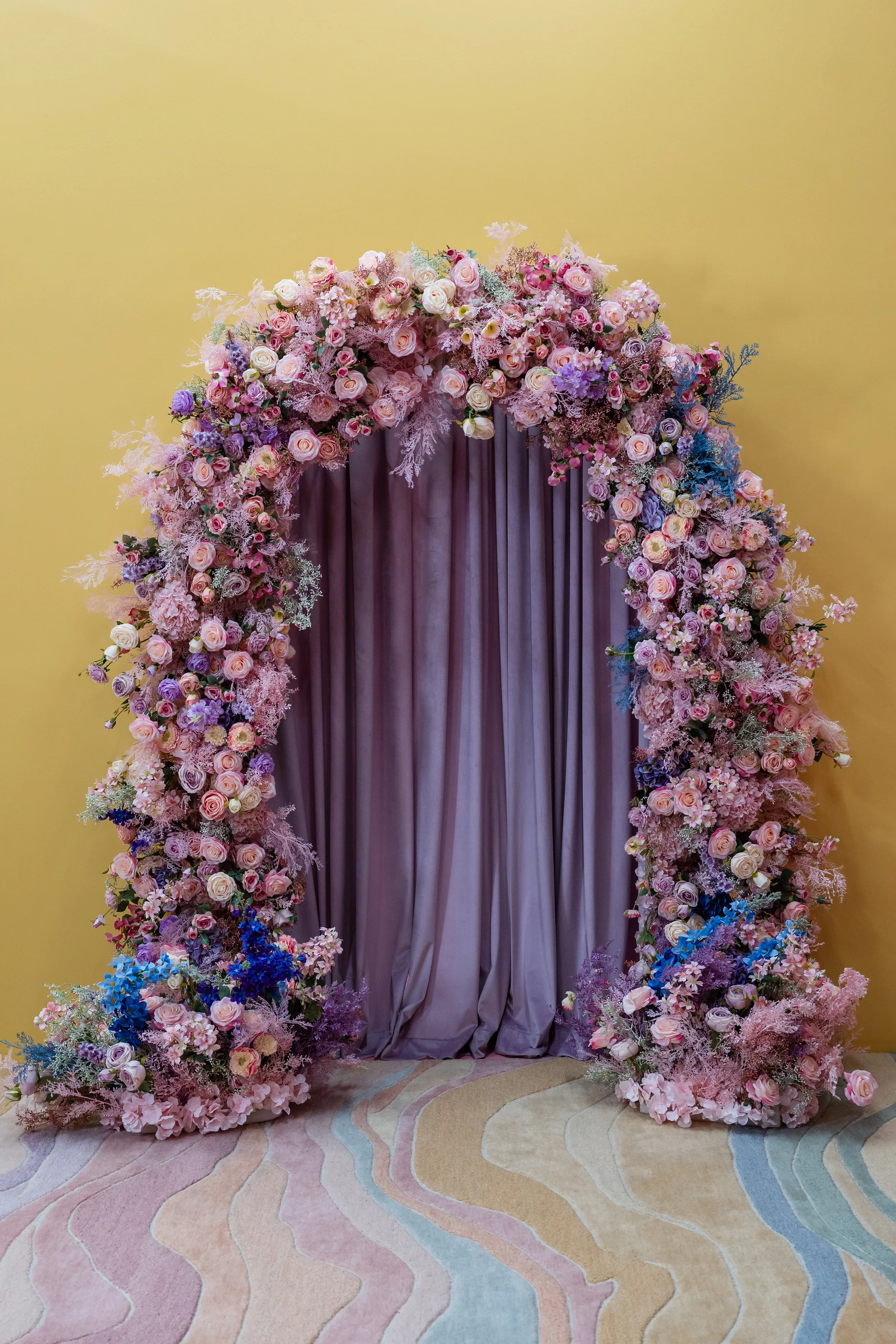 A floral arch with pink, purple, and white flowers and greenery, with a purple curtain background, on a pastel-colored patterned carpet against a yellow wall.