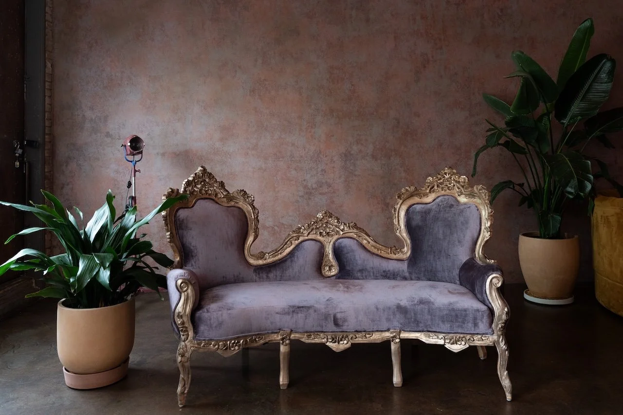 An ornate vintage sofa with dark purple velvet upholstery and carved gold detailing sits against a textured pink wall, flanked by large potted green plants.