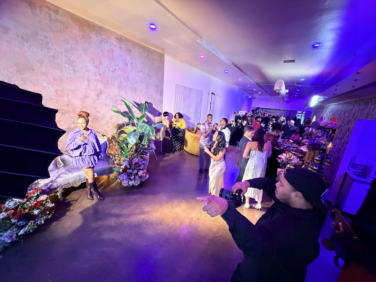 People dancing and mingling at an indoor celebration or party with colorful lighting, floral arrangements, and a buffet table in the background.