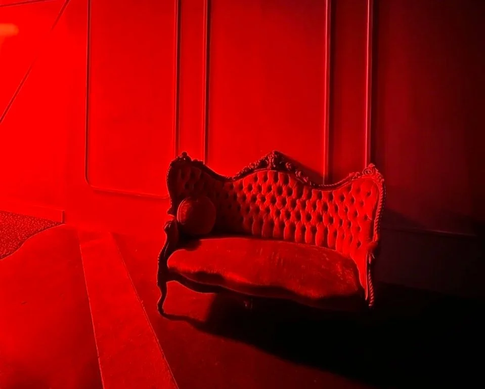 A vintage red velvet chaise lounge with tufted upholstery and ornate wood carvings, placed in a dimly lit room with red lighting.