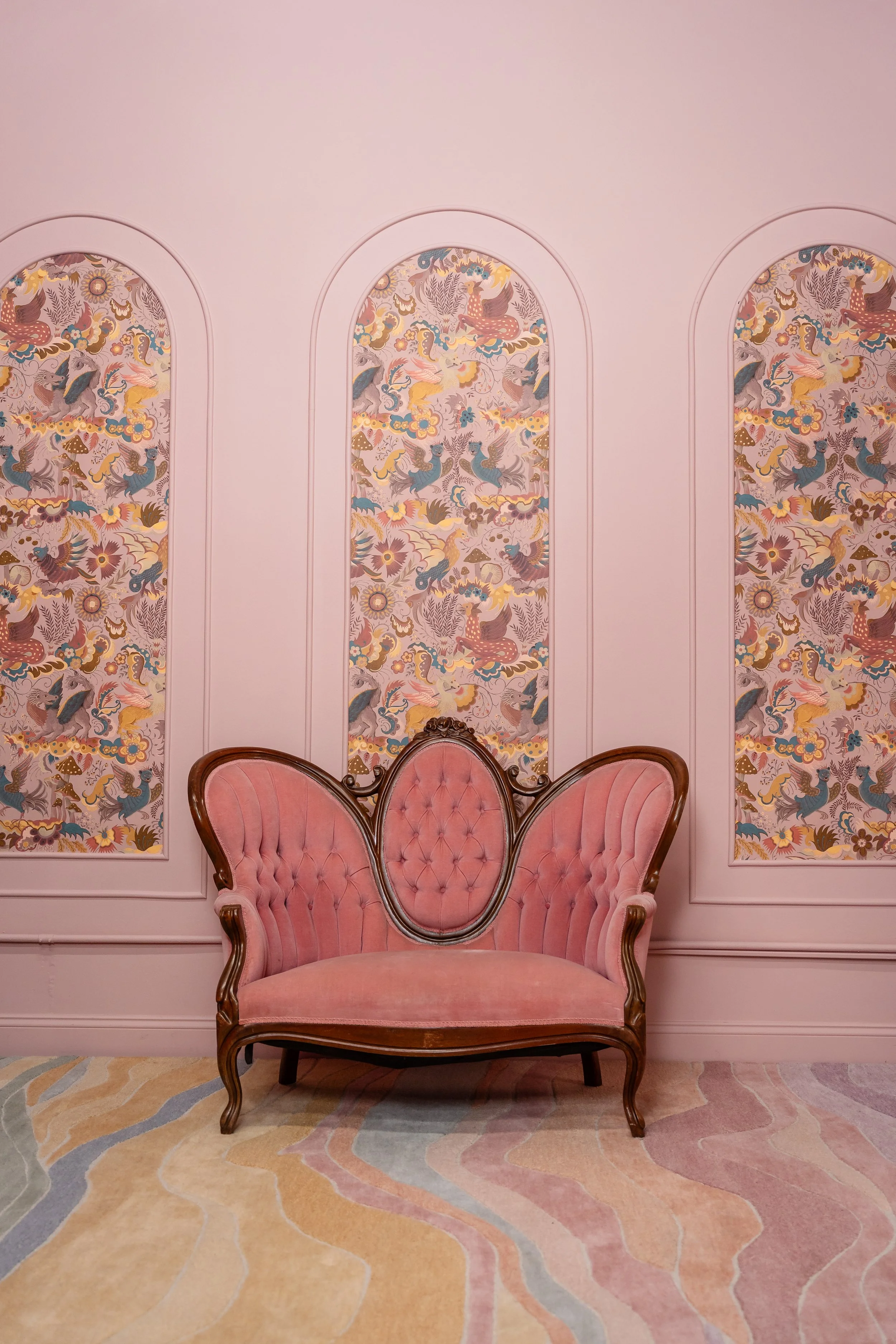 A vintage pink upholstered loveseat with carved wooden frame, set against a pink wall with three vertical decorative panels featuring a colorful dragon and floral design, and sitting on a multicolored abstract patterned carpet.