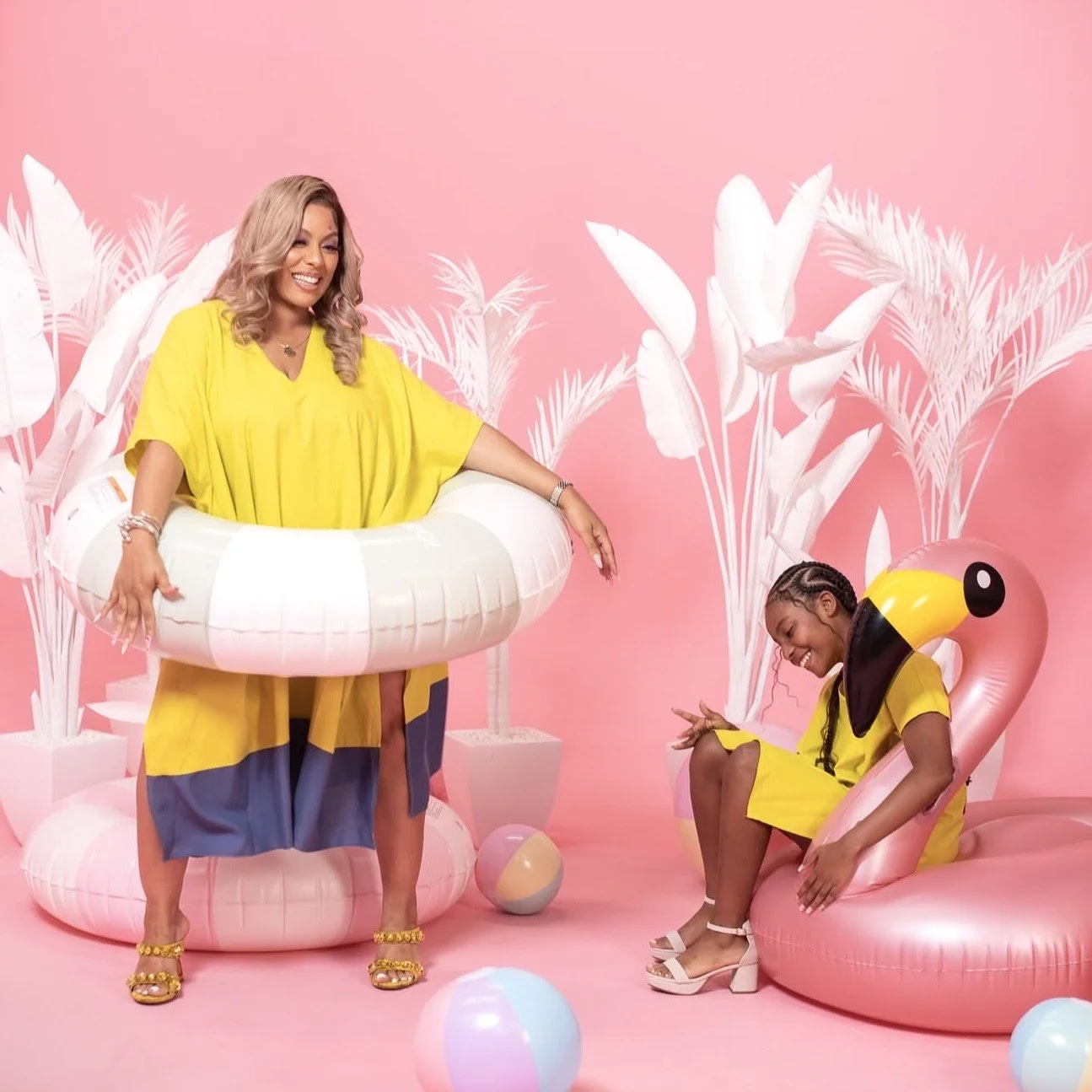 Two women in bright yellow outfits in a pink tropical-themed setting. One woman is sitting in a flamingo-shaped chair wearing cruise attire and the other is standing in a paddle floaty with a pink background, white tropical leaves, and beach balls ar