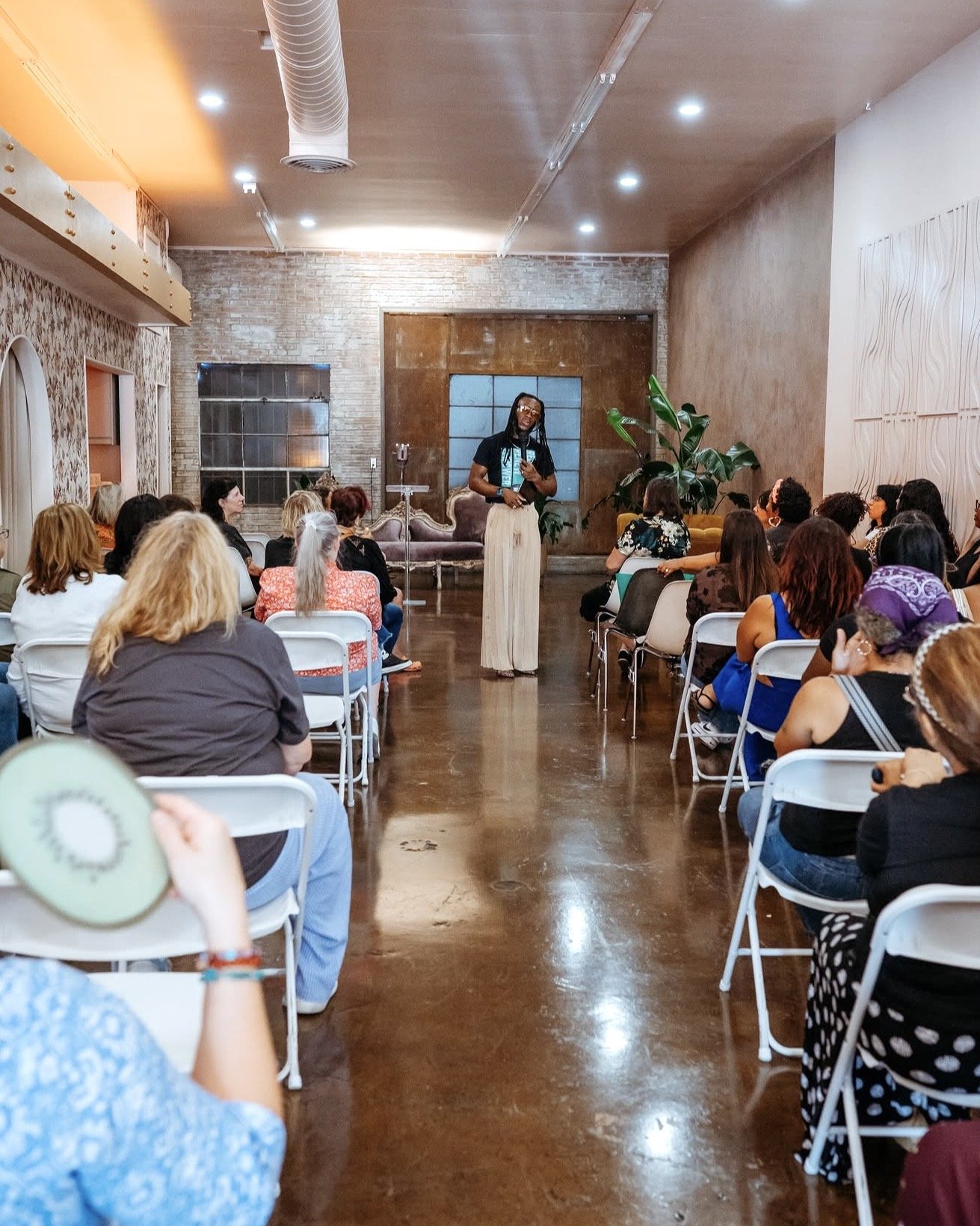 A woman standing and speaking in front of an audience in a room with exposed brick and wood walls, ceiling with recessed lighting, and a large plant. Event space Burbank