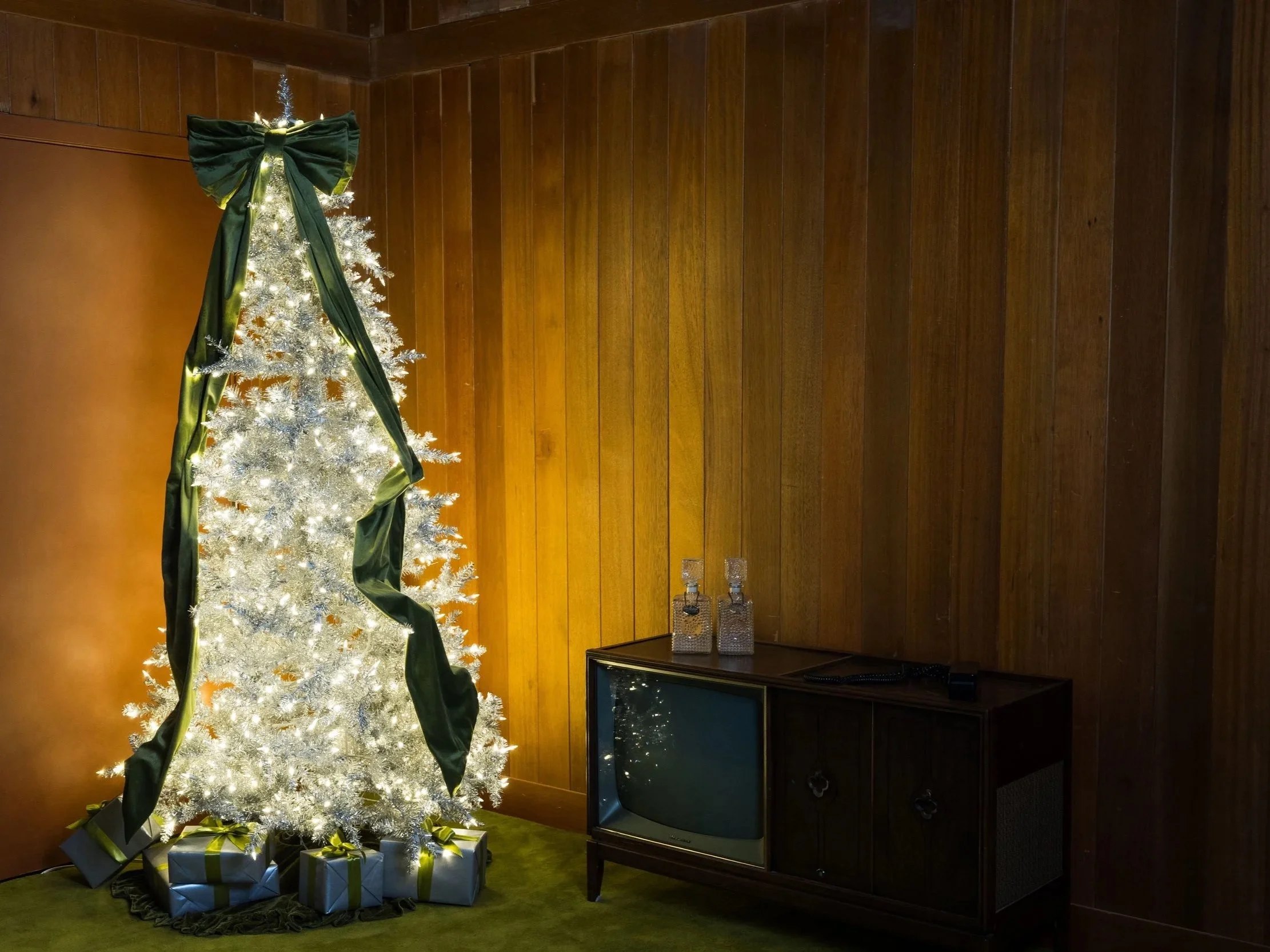 White Christmas tree decorated with lights and bows in a room with wood-paneled walls, surrounded by wrapped presents and an old-fashioned television.