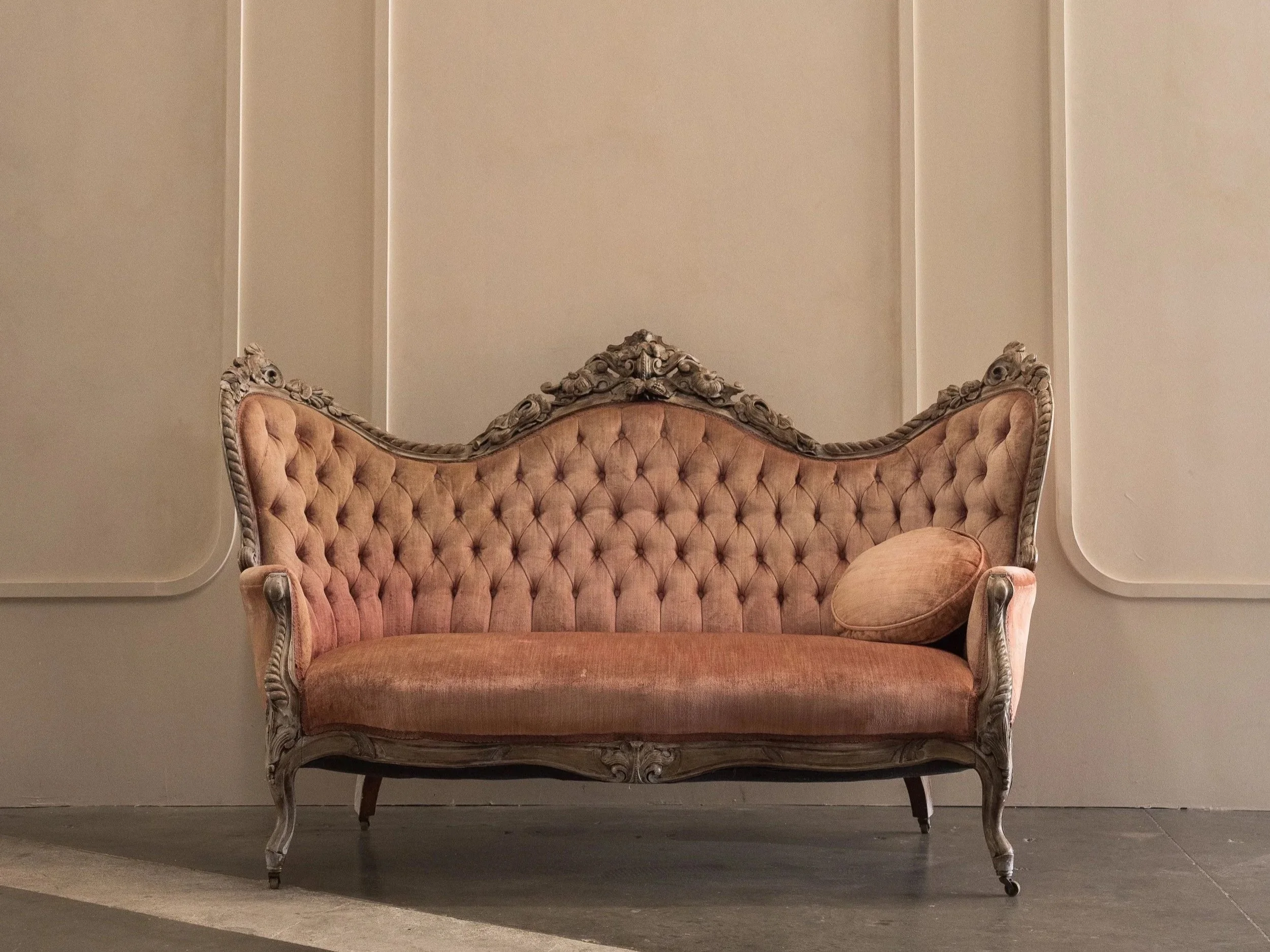 A vintage wooden framed sofa with pink velvet upholstery, tufted backrest, and a round bolster pillow against a black textured background.