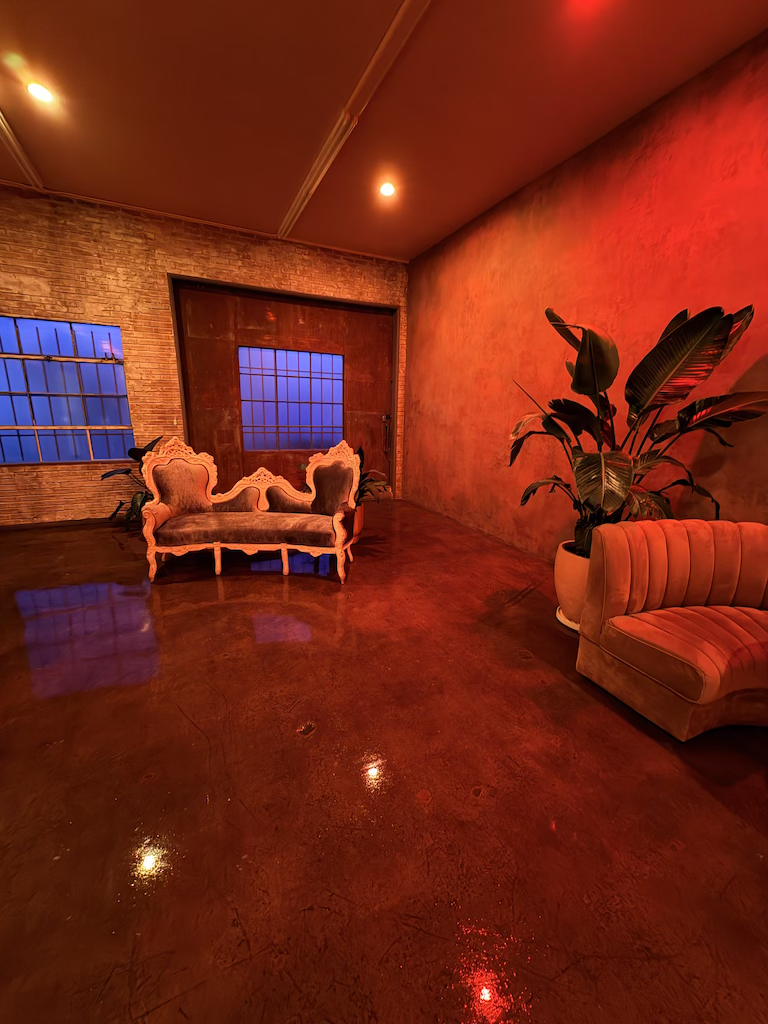 A dimly lit room with exposed brick and painted walls, a vintage sofa, a curved velvet chair, a large potted plant, and blue-lit windows.