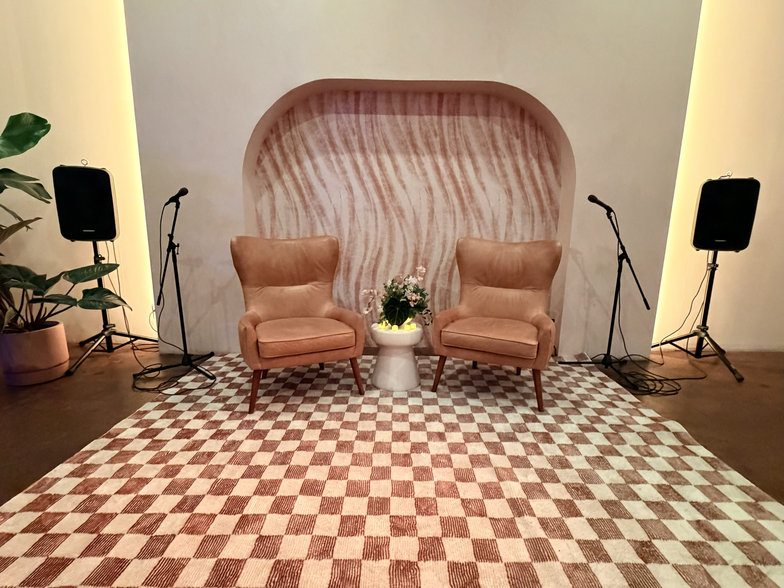 An indoor event setup with two light brown armchairs facing each other, a small round white table with a flower arrangement in the center, and a checkered rug on the floor. There are two microphones on stands and two large speakers on either side. Th