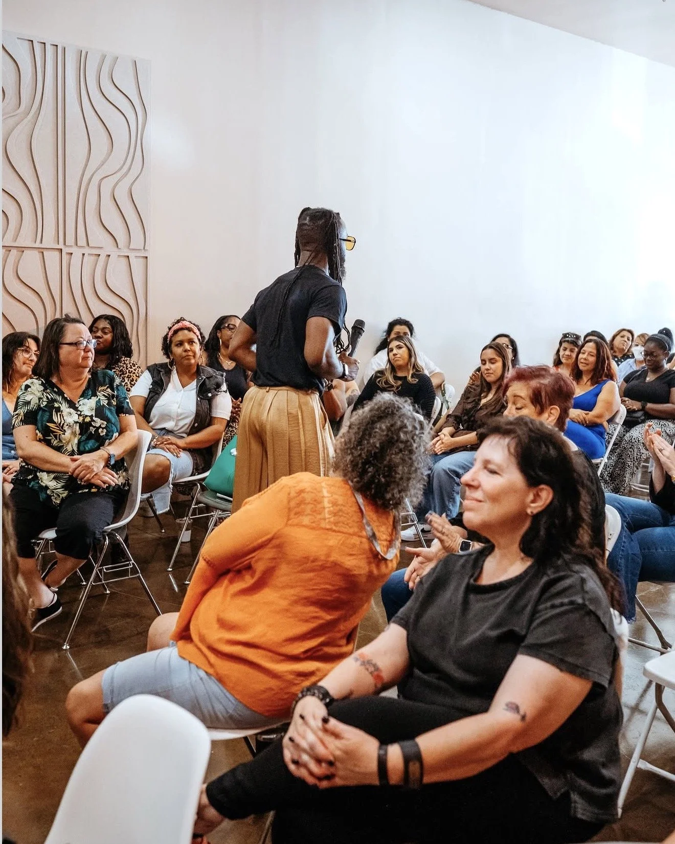 A group of women sitting in a room, listening to a woman standing with a microphone. The room has a modern design with decorative wall panels. Event Space Burbank