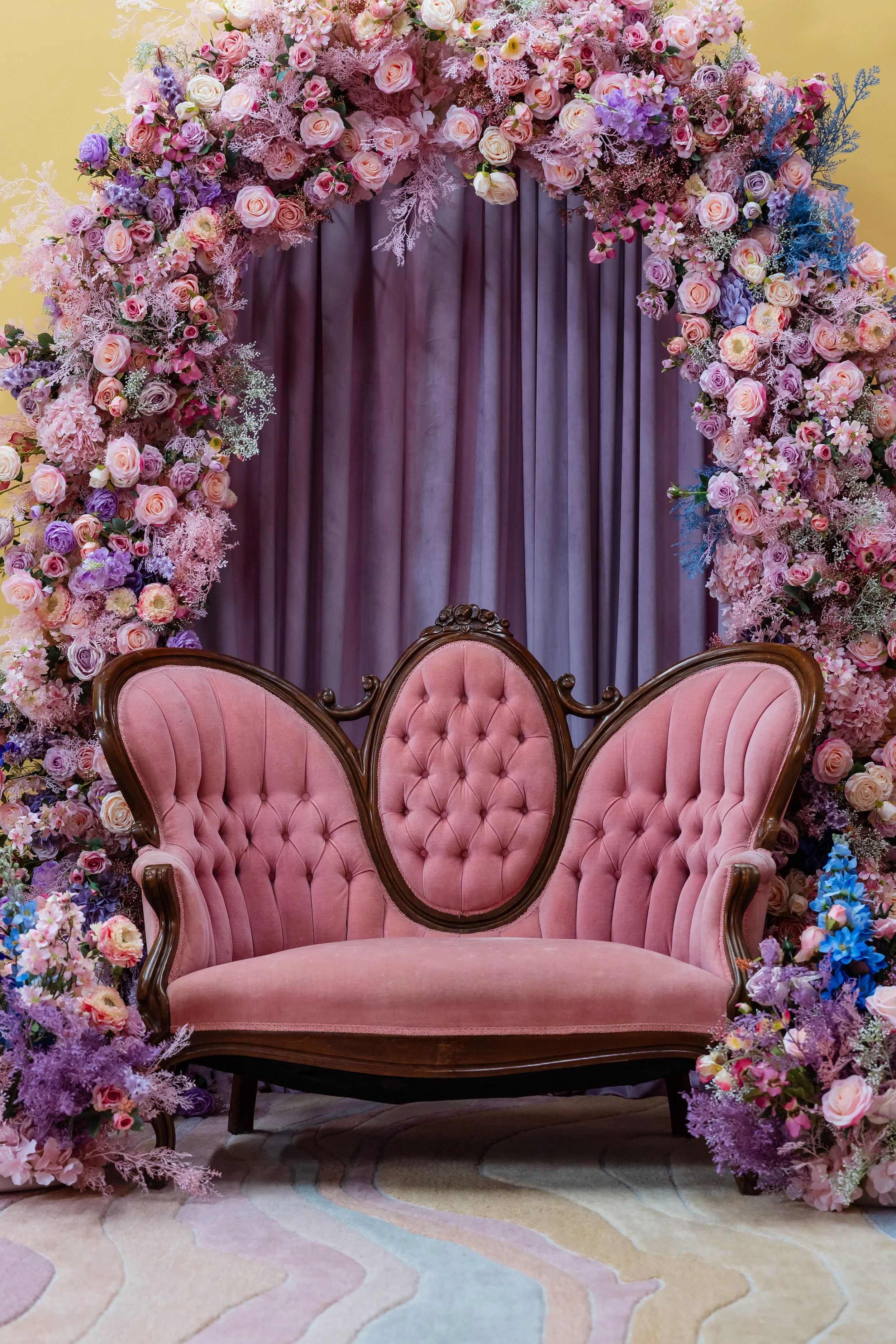 A vintage pink velvet tufted settee in front of purple curtains, surrounded by a large arch of pink, purple, and white flowers, including roses, hydrangeas, and other floral arrangements.