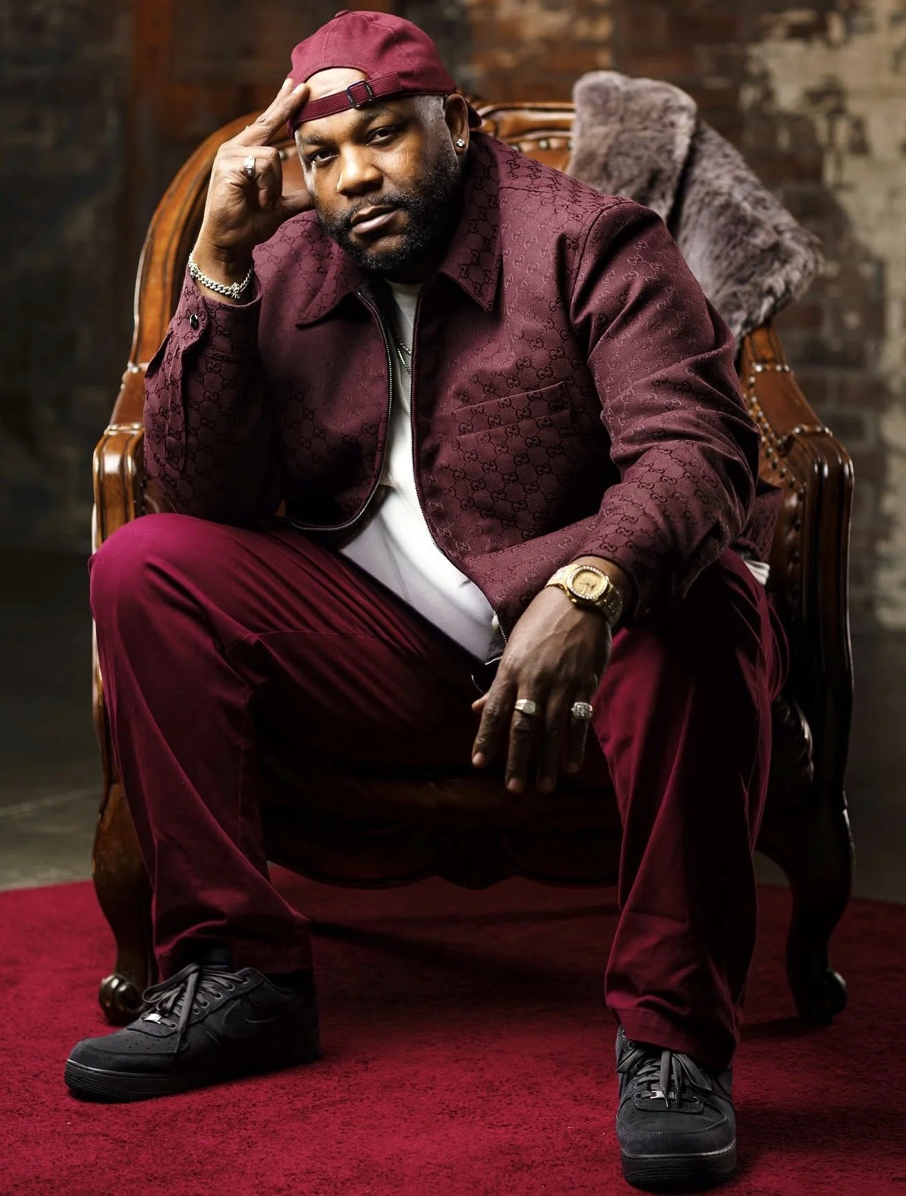 A man sitting on an ornately carved wooden chair with a fur throw pillow behind him. He is wearing a burgundy patterned jacket, matching burgundy pants, a maroon cap worn backwards, and black sneakers. He has a beard and is accessorized with rings, a