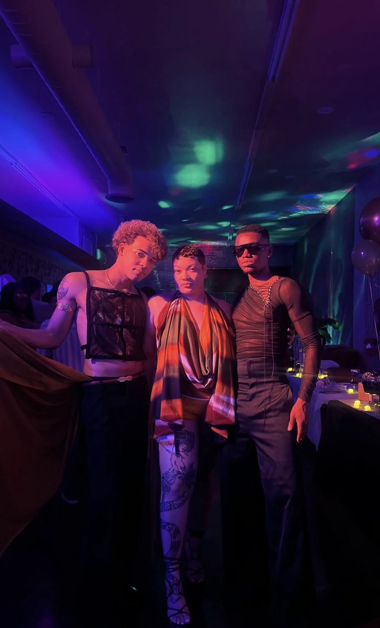 Three people posing at a nightclub with colorful lighting and balloons in the background. The person on the left has short curly hair, tattoos, and is wearing a black mesh outfit. The person in the middle has short hair and is wearing a multicolored 