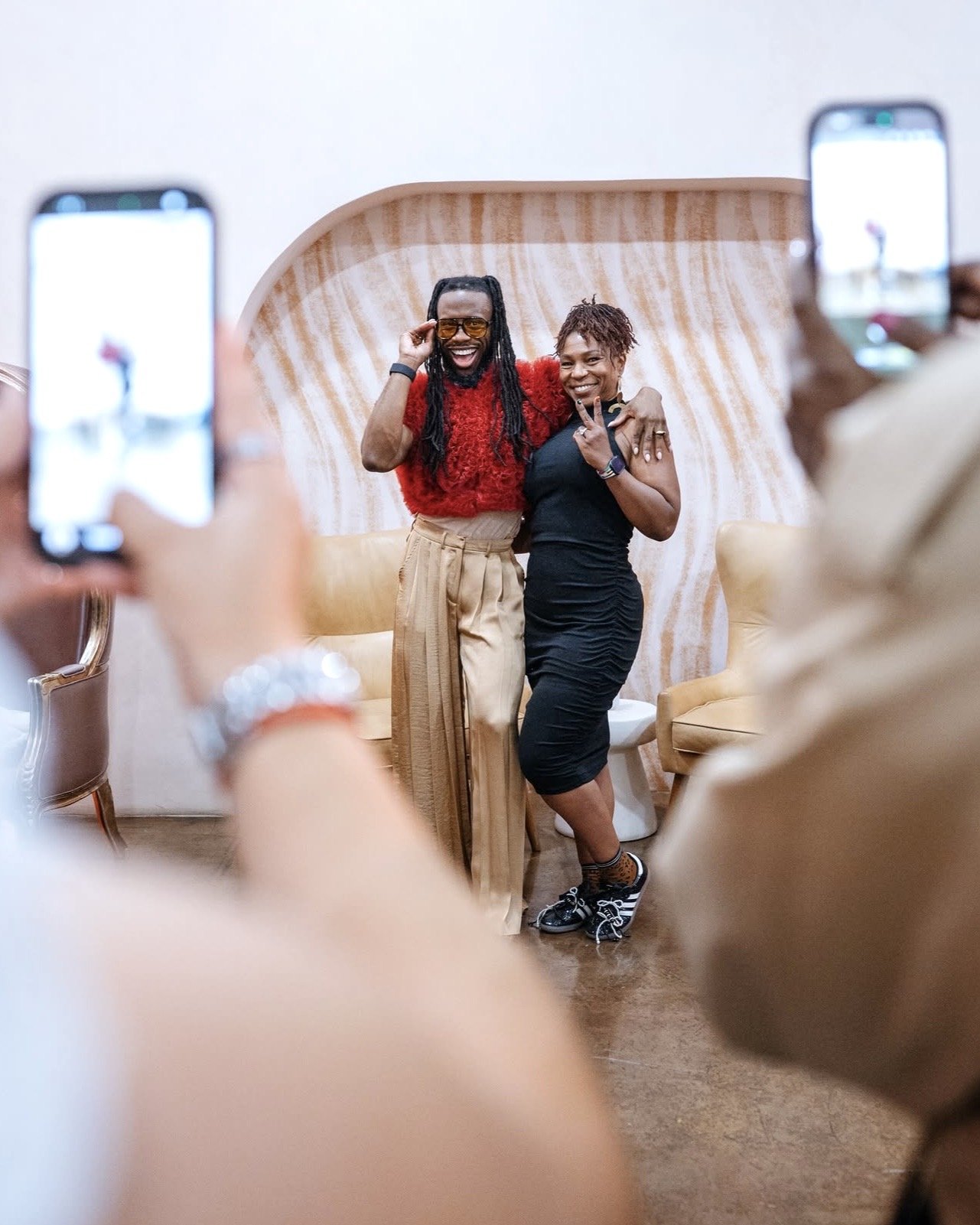 Two people standing close together, smiling and posing for photos indoors with a wooden wall background; one wearing a red top and beige pants, the other in a black dress. Event space Los Angeles.