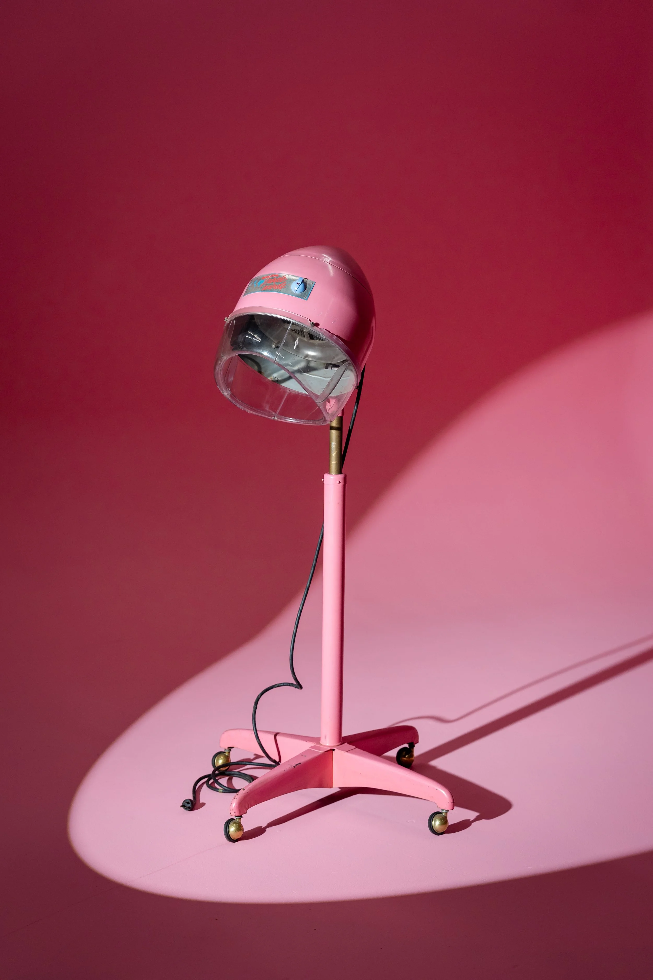 A vintage pink hairdryer on a pink stand with wheels, casting a shadow on a pink background.