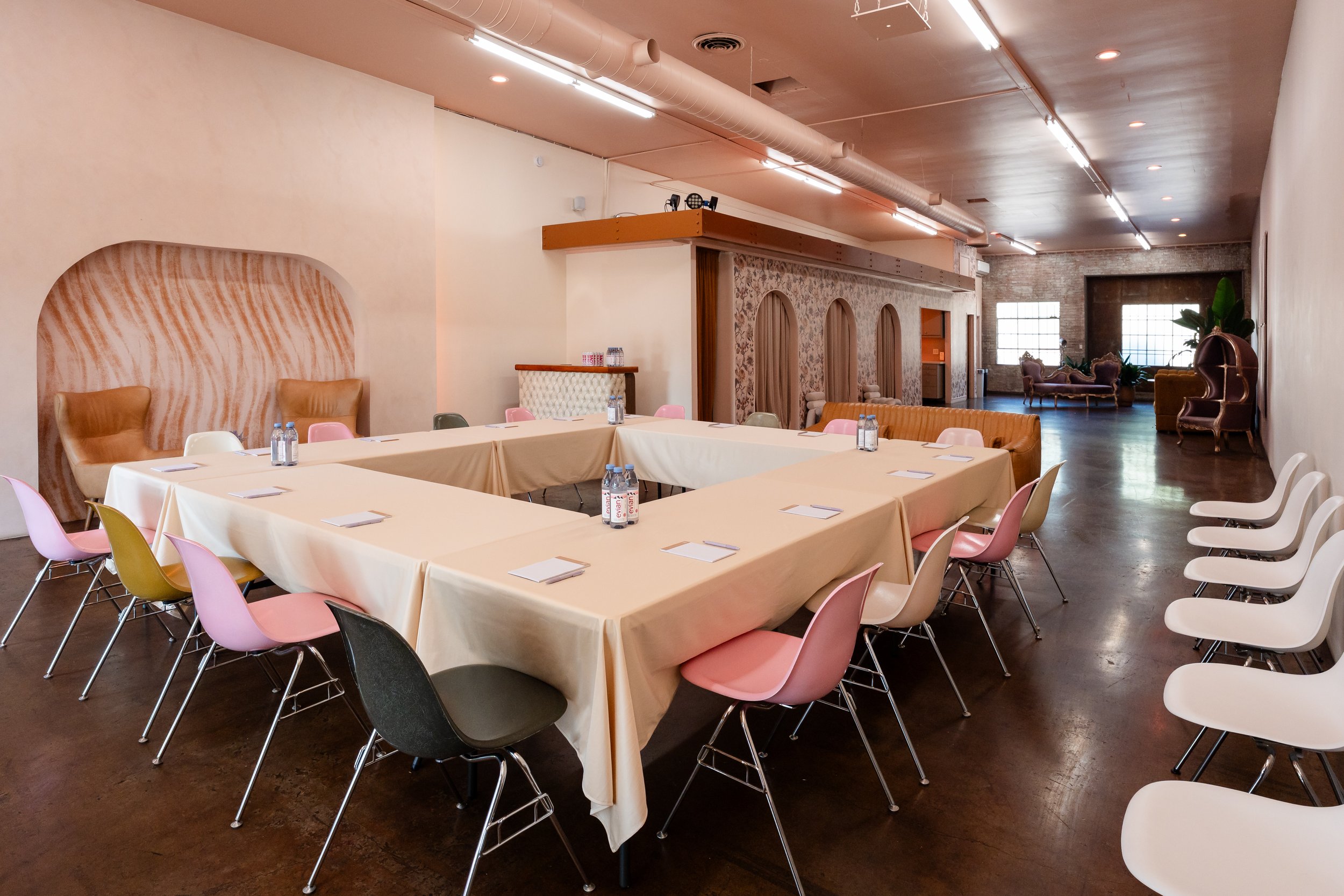 A spacious event or conference room with a large U-shaped arrangement of tables covered with beige tablecloths, surrounded by assorted pink, white, and black chairs. Bottled water is placed on the tables, and elegant chairs and couches are arranged a