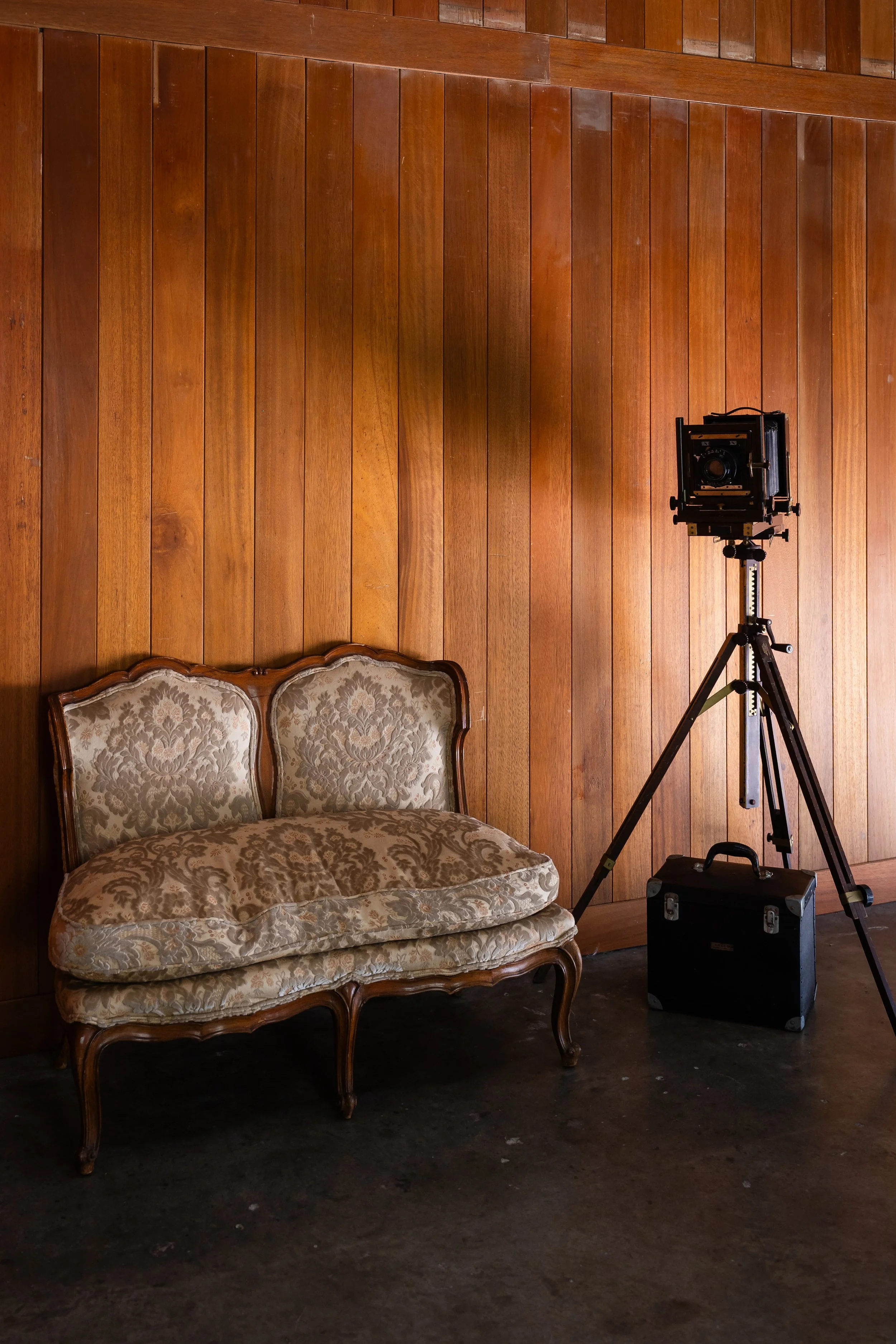 Vintage upholstered loveseat next to a professional camera on a tripod against a wood-paneled wall.