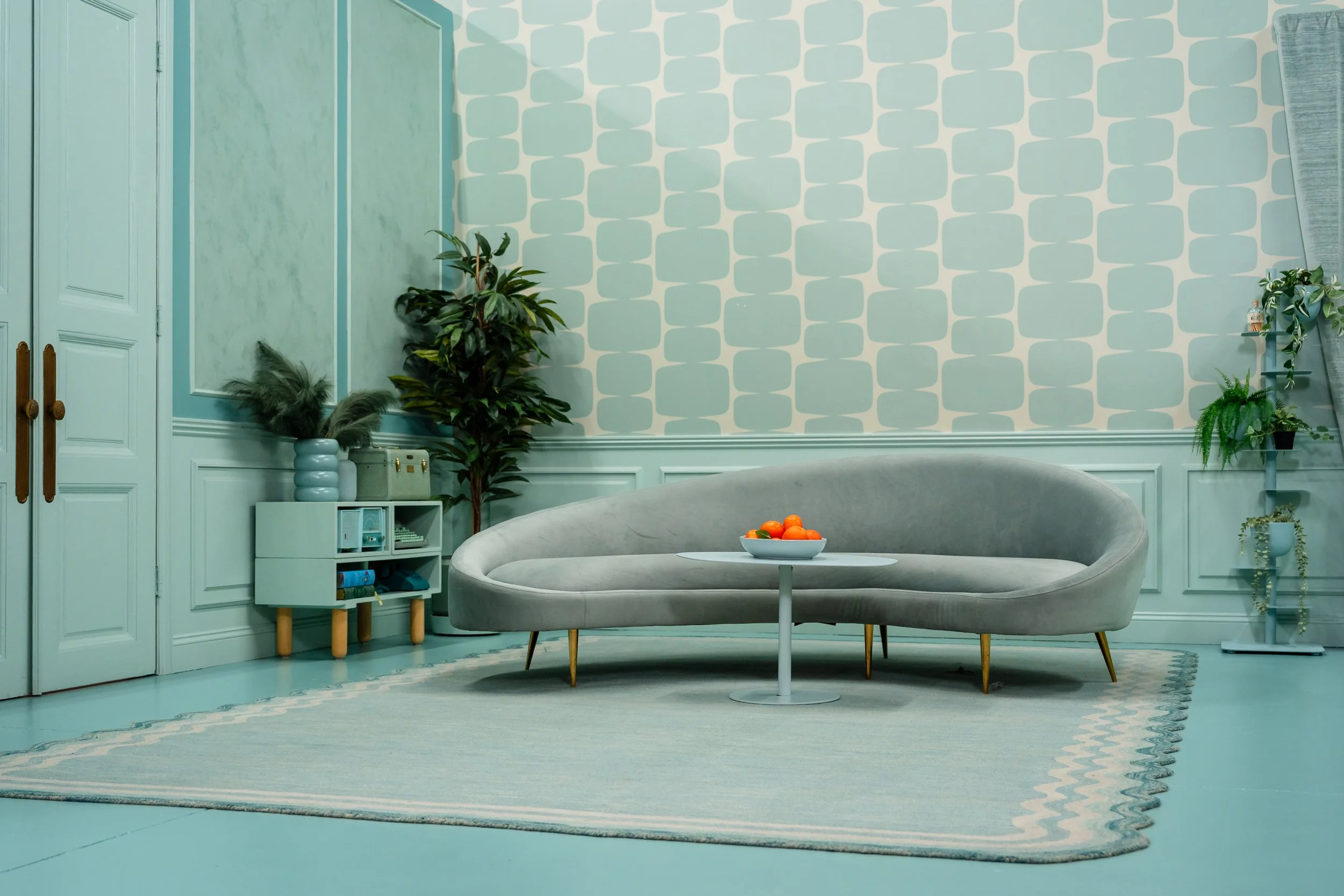 A modern living room with light teal walls, a gray curved sofa, a small round white table with a bowl of oranges, a patterned rug, potted plants, and shelves with decorative items.