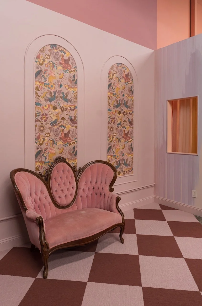 Vintage pink velvet tufted loveseat with ornate wood trim in a pastel-colored room. Two arched wall panels with floral and mythical creature patterned wallpaper. Checkerboard floor in pink and beige.