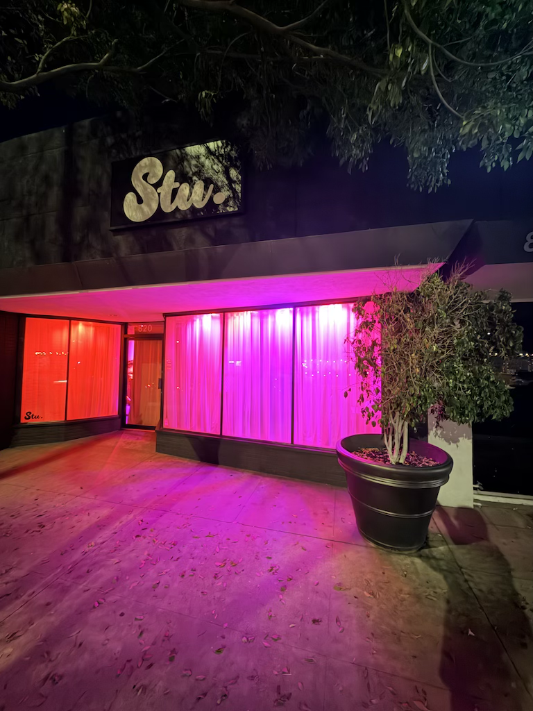 Night view of a storefront with bright pink and red neon lighting illuminating the windows and a potted tree outside. The sign above reads 'Stw.'