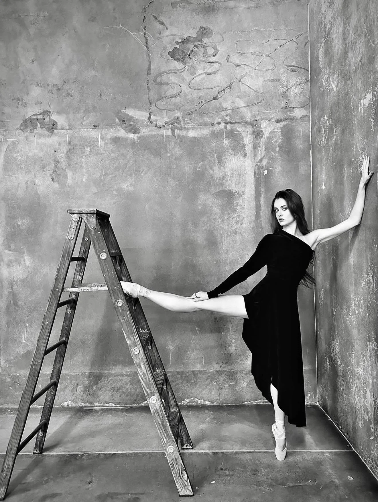 A ballerina in a black dress in an old, industrial-style room with concrete walls. She is balancing on one leg with her hand on the wall and the other arm reaching out. A wooden ladder is in front of her, and she is wearing ballet shoes.
